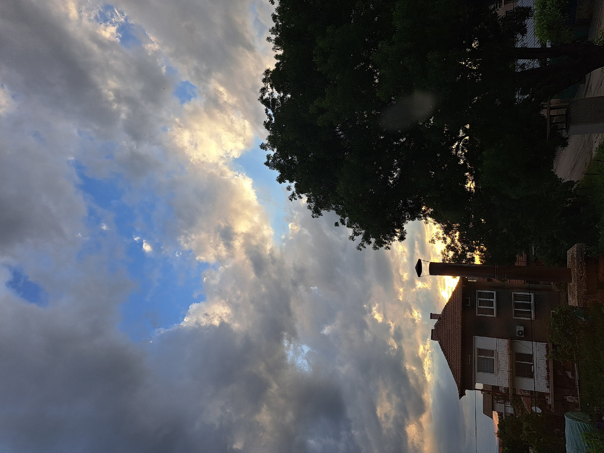 The image, taken with a vertical orientation, captures an outdoor scene at either sunrise or sunset, characterized by dramatic lighting and cloud formations.
The sky dominates the upper and left portions of the frame (when viewed correctly oriented, it would be the upper half). It features a dynamic mix of dark grey, heavy clouds and bright, golden-white clouds illuminated by the low sun. Patches of clear, vibrant blue sky are visible between the clouds.
In the lower right, a two-story residential house is partially visible. It has a reddish-brown tiled roof and light-colored walls with multiple windows, some featuring white frames and decorative white shutters. An air conditioning unit is mounted on the second floor. A tall, dark chimney or pipe extends upwards from near the house, silhouetted against the bright sky.
A large, dense-foliaged tree, dark green in color, occupies the upper right side of the image, its branches reaching towards the sky and partially obscuring the view. The strong backlight from the sun creates silhouettes of the tree and parts of the house. No people or specific activities are depicted. The overall impression is one of a quiet, atmospheric moment at dusk or dawn. There is no visible text.