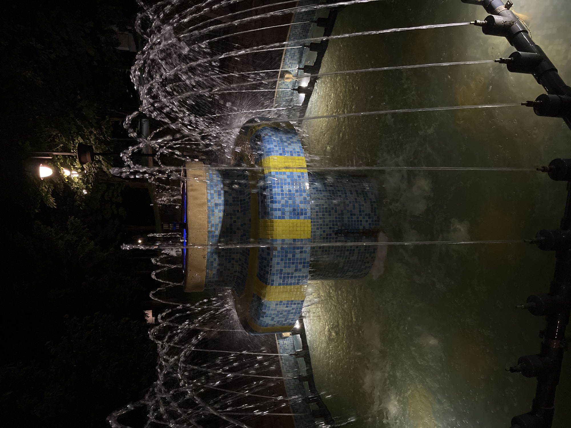 This nighttime image depicts an illuminated, multi-tiered public fountain situated in Veliki Preslav, Bulgaria. The fountain's structure is adorned with mosaic tiles, predominantly blue, featuring a distinct yellow horizontal band on its middle tier. Water actively sprays from numerous nozzles, forming multiple high arcs and vertical jets that cascade into a circular lower basin. The water in the basin is illuminated by submerged lights, giving it a greenish-yellow hue and creating visible agitation. Along the rim of the lower basin, a dark-colored pipe system is visible, equipped with multiple individual nozzles that project streams of water outwards. The uppermost tier of the fountain emits a blue light. The background is composed of dense, dark foliage, likely trees, silhouetted against the night sky. Two streetlights with warm, yellow-toned illumination are visible above the treeline, contributing to the ambient light. No individuals or animals are present in the scene.