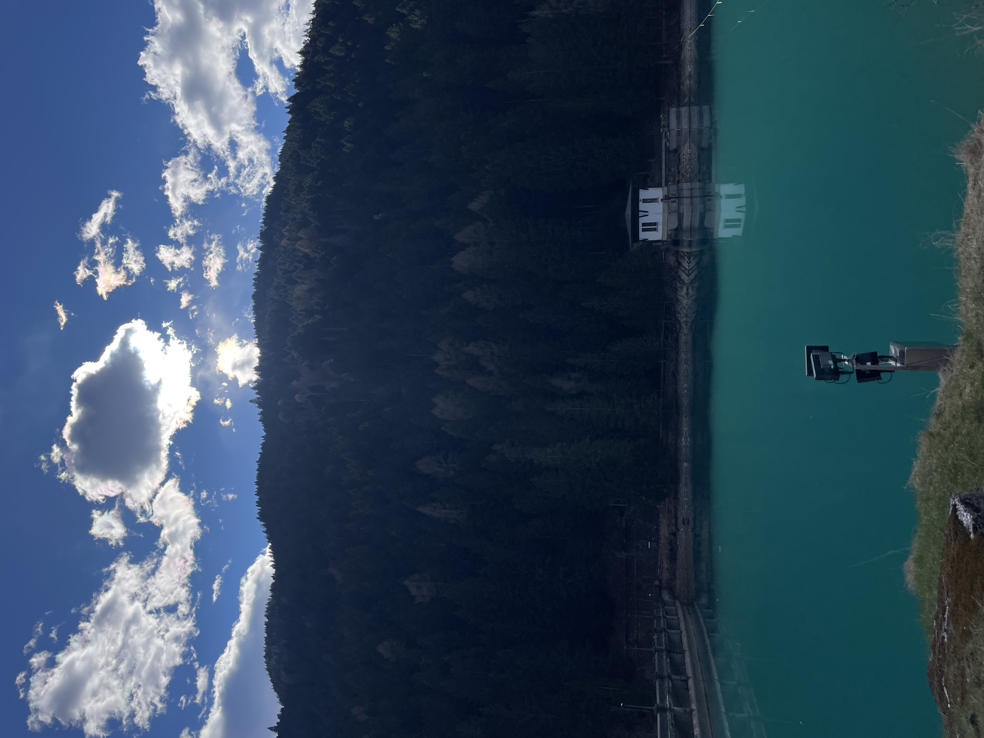 The image depicts a reservoir, likely in the mountains near Auronzo di Cadore, Italy. A forested hillside runs along the edge of the water. A small white building sits on a platform extending into the reservoir. A metal structure stands on the bank near the reservoir. The sky is visible to the left and shows blue sky with white clouds.