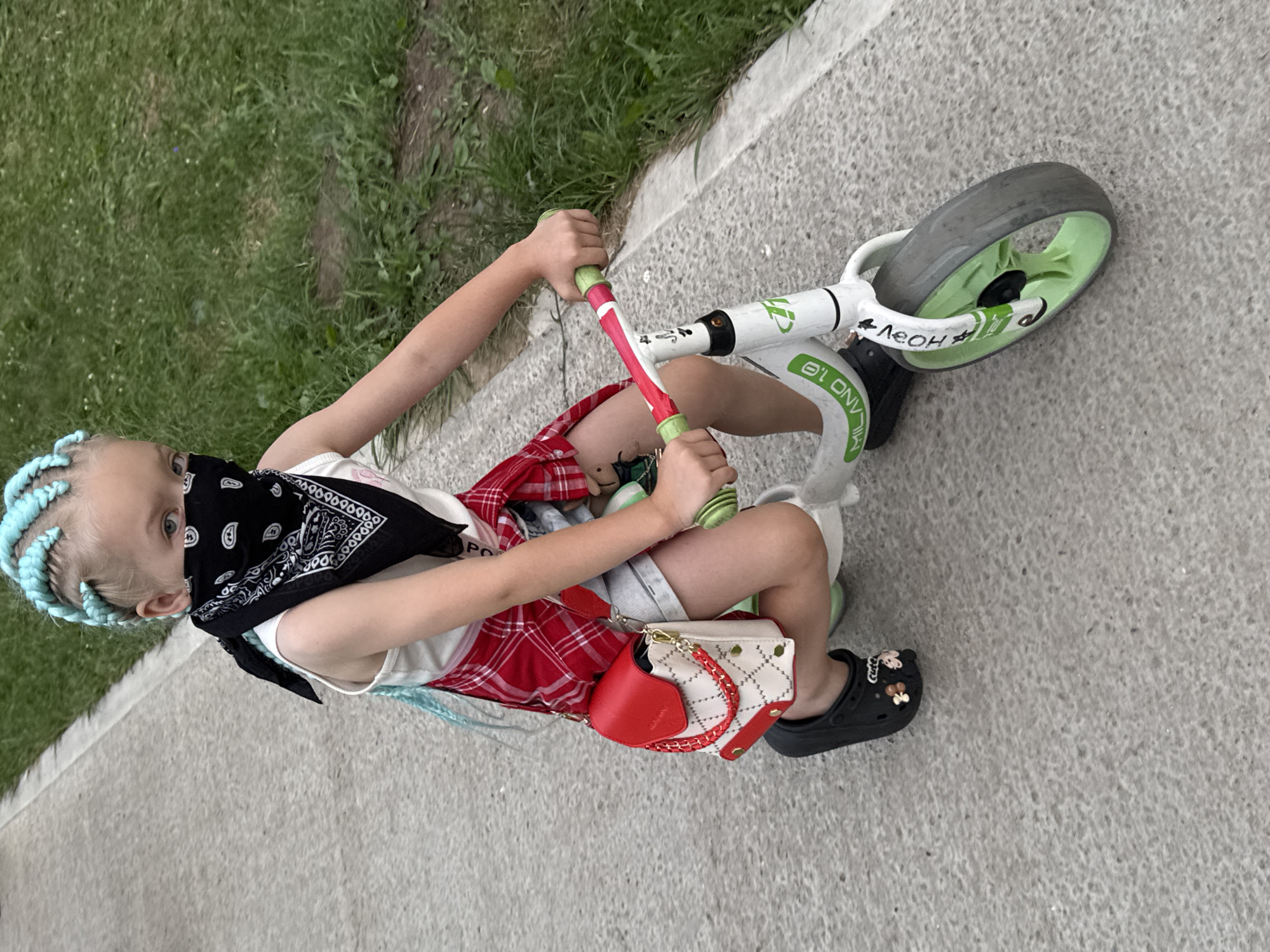 A young girl with blue braided hair and a black bandana covering her mouth and nose rides a white balance bike with green accents. She is wearing a white tank top, a red and white plaid shirt tied around her waist, and denim shorts. A small red and white purse with a gold chain is slung over her shoulder. She is wearing black Crocs with flower and bear charms. The bike has "MILANO 1.0" and other green lettering on its frame. The girl is on a paved path next to a grassy area. The setting appears to be a park or recreational area, possibly in Suzdal, Russia, given the architectural style of the buildings in the background and the overall aesthetic.