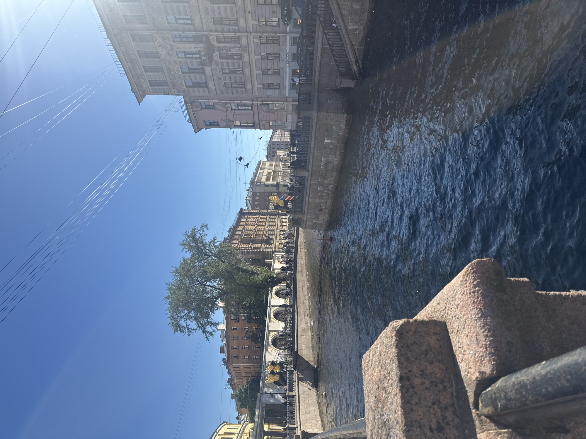 This is a daytime, eye-level, medium shot of a canal in Saint Petersburg, Russia, likely taken from a bridge. The canal's water is a dark blue, reflecting the clear, bright blue sky with scattered white contrails from aircraft. On the right bank of the canal are several multi-story buildings with ornate architecture; one building is pinkish, and another is a light gray stone. The buildings feature multiple windows and decorative elements. Along the canal's edge, there is a stone embankment topped with a dark metal railing. People are visible walking along the embankment and on a bridge across the canal. On the left side of the canal, there's a distinct architectural structure with arches, a gilded statue of a winged figure, and bicycles parked nearby. A large tree with green foliage stands behind this structure. The foreground of the image shows a close-up view of a granite parapet and a dark metal handrail. Sunlight creates reflections on the water and casts shadows. A red buoy is visible in the canal.