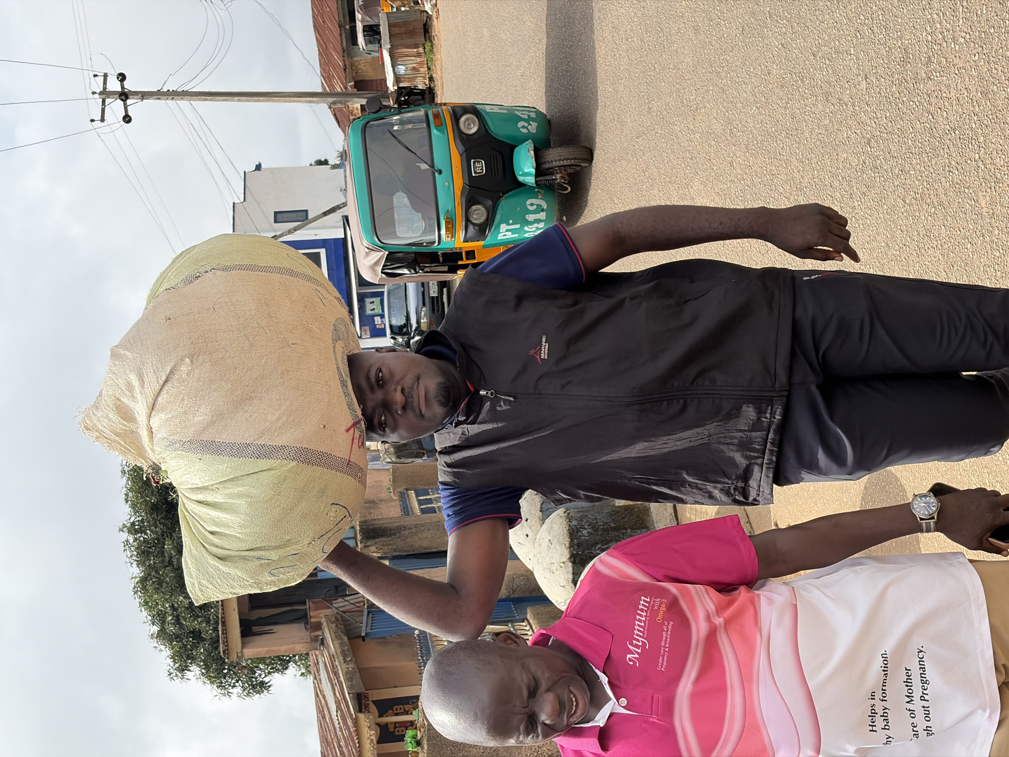 In Jos, Nigeria, a man is captured in a medium shot carrying a large, light-colored sack of goods on his head. He is wearing a dark vest over a blue and red shirt and black pants, with a watch on his left wrist. Beside him, to his left and slightly in front, another man is visible from the chest down, wearing a pink and white patterned polo shirt with text that reads "Mymum" and other details related to pregnancy and baby care, and a watch on his left wrist. He appears to be looking down and to his left. In the background, a green and yellow motorized tricycle, commonly known as a "Keke Napep" or "Marwa," is parked on the right side of the paved road. Other vehicles and buildings with corrugated iron roofs are also present, suggesting an urban or semi-urban setting. The sky is overcast. The scene depicts everyday life in a Nigerian city.
