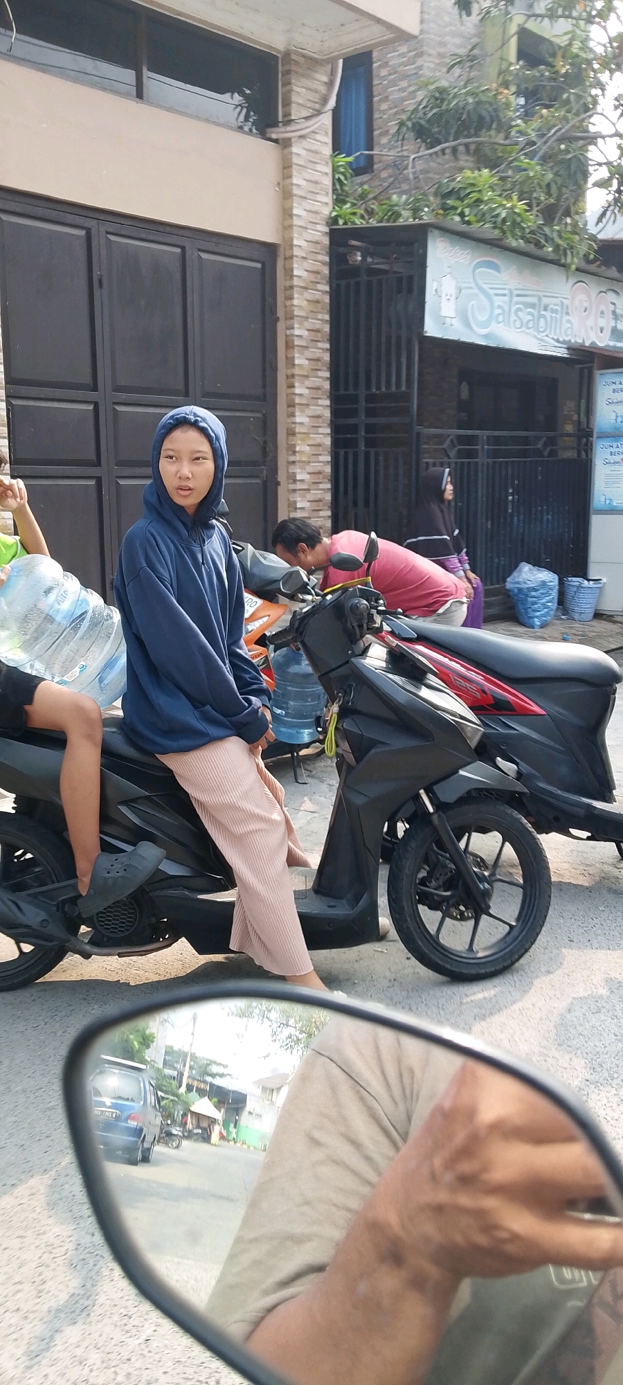 In Satriamekar, Indonesia, a young individual wearing a blue hoodie and pink pants sits on a black motorcycle. To their left, a large plastic water bottle is visible. Behind them, a person in a pink shirt is leaning over another motorcycle. Further in the background, a woman wearing a dark hijab and purple clothing sits on a motorcycle near a building with a sign that reads "Salsabilah Ro". The scene appears to be on a street in front of shops or residences, with a building facade and some vegetation visible. A motorcycle's side mirror in the foreground reflects a blue car and more of the street scene.