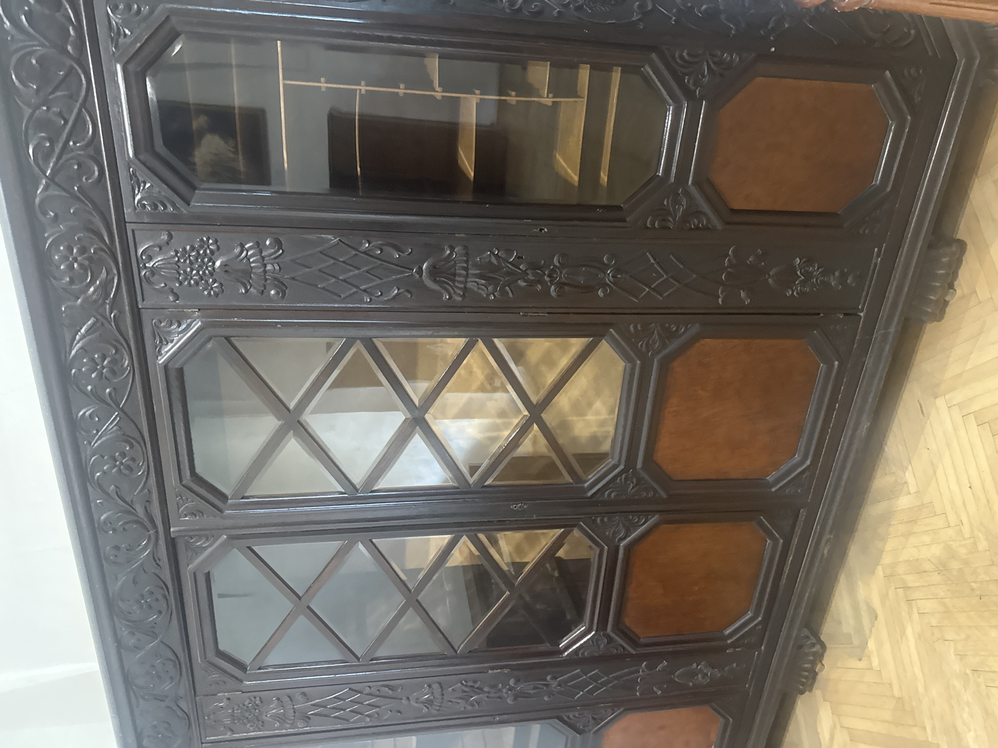 The image depicts a dark, ornate wooden cabinet with glass panels and decorative carvings. The cabinet has multiple tiers of glass-paneled doors, some with a diamond pattern, and solid wood panels arranged in octagonal shapes. The background is blurred, but the floor appears to be wooden with a herringbone pattern. The scene appears to be indoors.