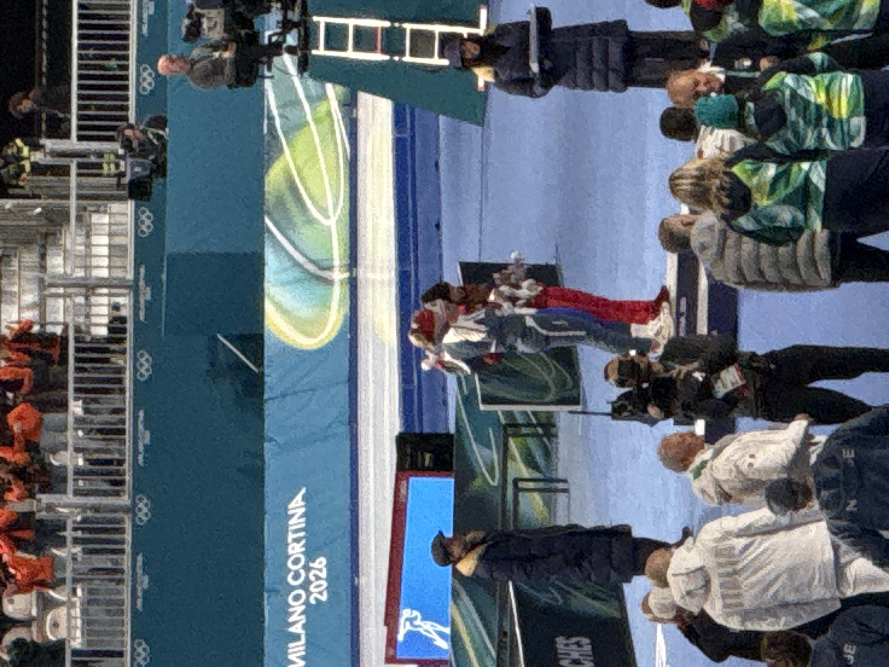 Attendees and athletes stand around a banner displaying the "Milano Cortina 2026" logo. Several individuals are wearing winter jackets. A few photographers are present, capturing the moment. The scene seems to be taking place in an outdoor area with a staircase visible in the background. The overall setting suggests an event or ceremony related to the 2026 Milano Cortina Olympics.