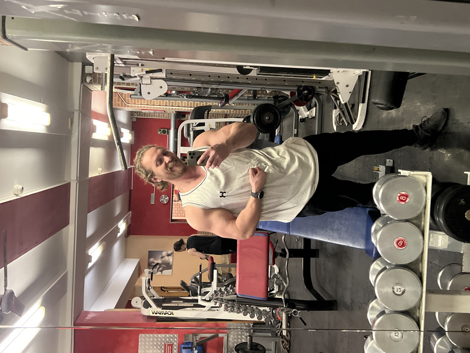 The scene depicts the interior of a fitness gym in Saint Petersburg, Russia, captured as a mirror selfie. A muscular male individual with light brown, curly hair, a beard, and a white Under Armour tank top stands facing a large mirror, holding a black smartphone in his right hand to take the photograph. He is smiling and has his left arm bent, flexing his bicep, with a dark smartwatch visible on his left wrist. He wears black athletic pants and dark shoes.
Reflected in the mirror, behind the primary subject, is the gym's interior. The ceiling features red and white striped panels with fluorescent lighting. The walls are predominantly red. Various gym equipment is visible: a rack of dumbbells in the foreground with weights including 15kg, 12kg, and 8kg (identified by red numerical markings). Behind the subject, a blue and red adjustable workout bench is positioned. Further in the background, reflected equipment includes a multi-station weight machine, a cable crossover machine, and various barbells and weight plates.
A second individual, likely female, with dark hair tied back and wearing a dark top, is partially visible in the reflection, standing near a piece of cardio equipment or a machine in the far background, facing away from the mirror. A framed black and white image, possibly a fitness-related poster, hangs on the wall behind this second person. A white sign with red lettering, likely in Russian, is also visible on the red wall.
