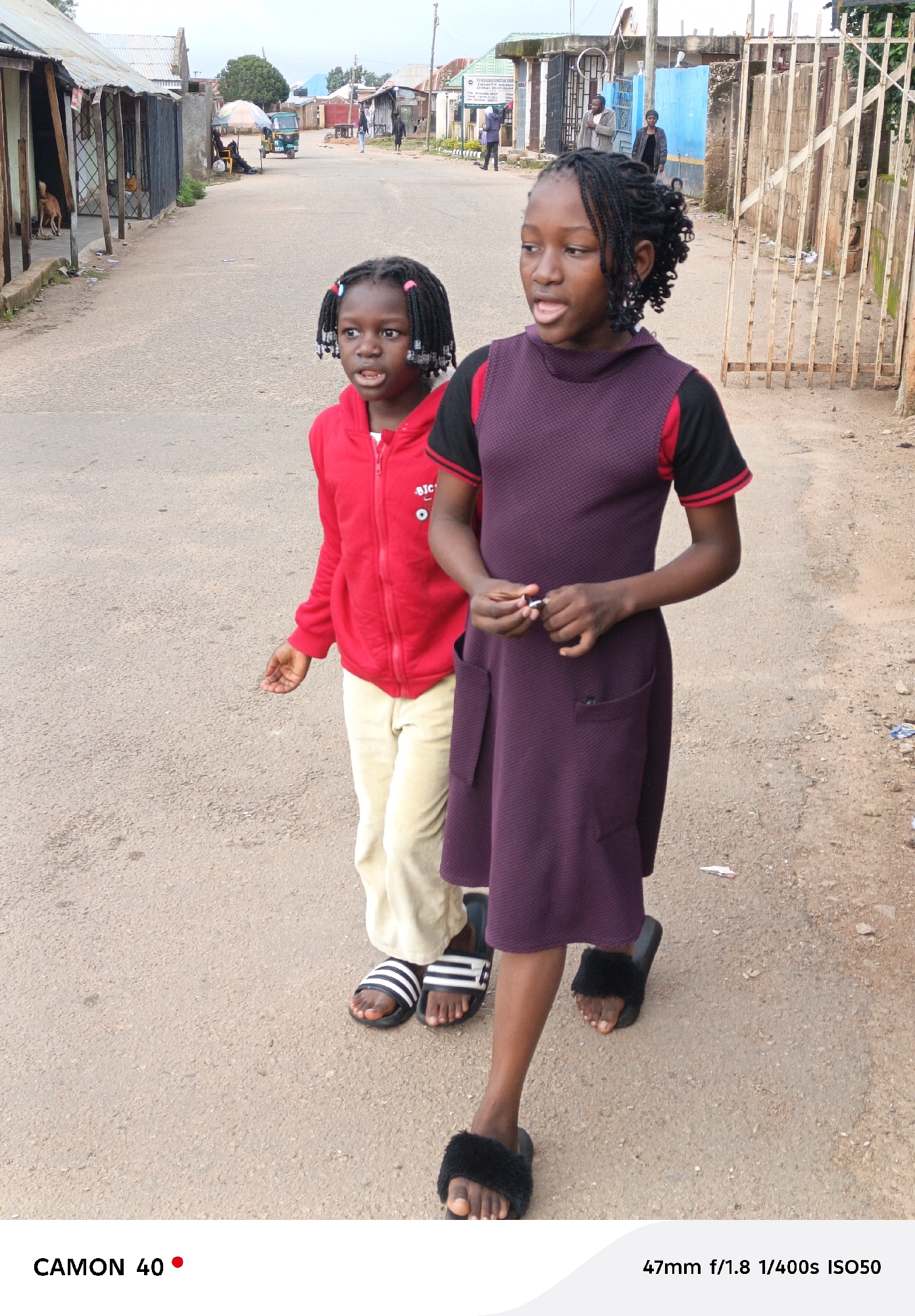 Two young Black girls, one wearing a red hooded jacket and cream-colored pants, the other a purple and black dress, walk along a paved road in Jos, Nigeria. The girl in the red jacket is on the left, and her face is turned slightly to the left, with her mouth open. The girl in the dress is on the right, facing forward, and appears to be holding something small and white in her hands. Both girls are wearing sandals. The background shows a street with buildings, a metal gate, and trees. The overall lighting suggests it is daytime.