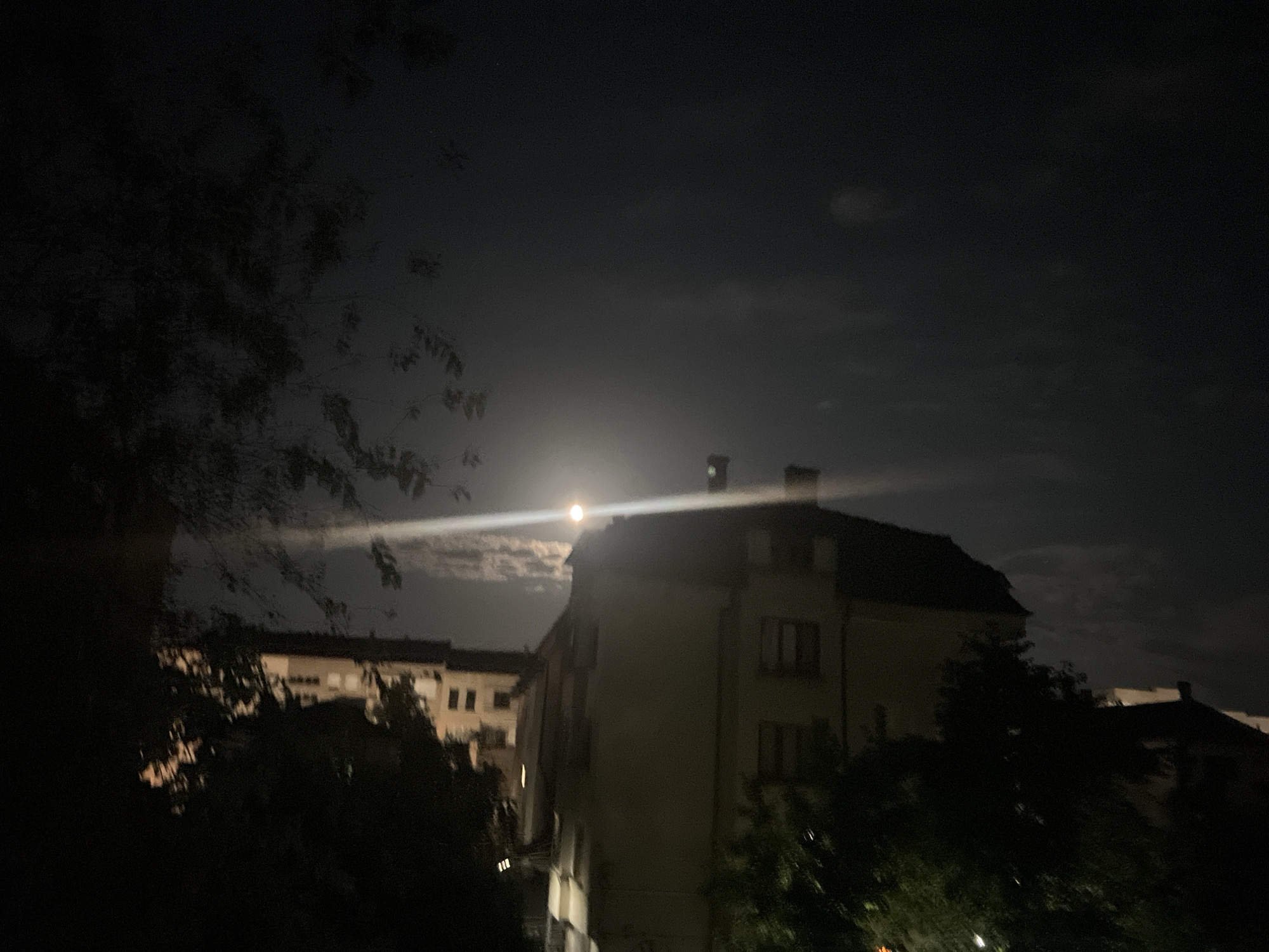 The image depicts a nighttime scene in the city of Blagoevgrad, Bulgaria, dominated by a dimly lit sky and silhouetted buildings. The moon is visible, partially obscured by clouds, and a bright beam of light, possibly from a streetlamp or a camera artifact, crosses the sky. In the foreground, dark foliage obscures the lower part of the frame. Behind the foliage, several buildings with visible windows and architectural details are present, some appearing to be residential or office structures. The overall ambiance is dark and somewhat atmospheric, characteristic of a night photograph taken with limited light.
