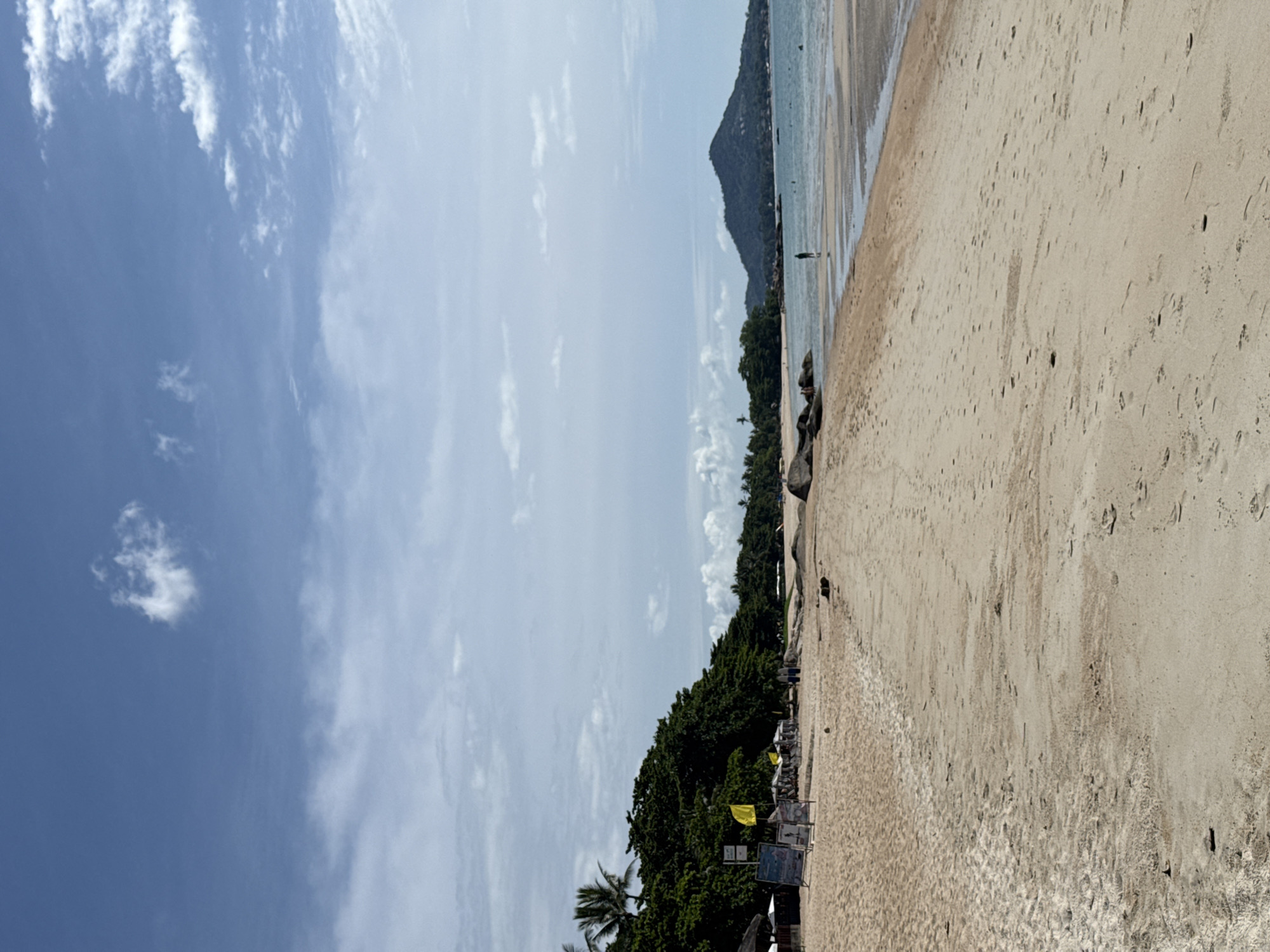 A wide shot captures a serene beach scene under a partly cloudy blue sky. The foreground is dominated by light brown sand with numerous footprints. To the left, a line of lush green trees borders the sandy expanse, with a few umbrellas and signs visible beneath them, suggesting a resort or dining area. A solitary yellow flag stands prominently among the trees. Further back, large rocks are scattered along the shoreline. The sea is calm and a pale blue, with a distant mountainous island forming the horizon. A single person stands in the shallow water, facing away from the camera, and a few other figures are visible near the water's edge.