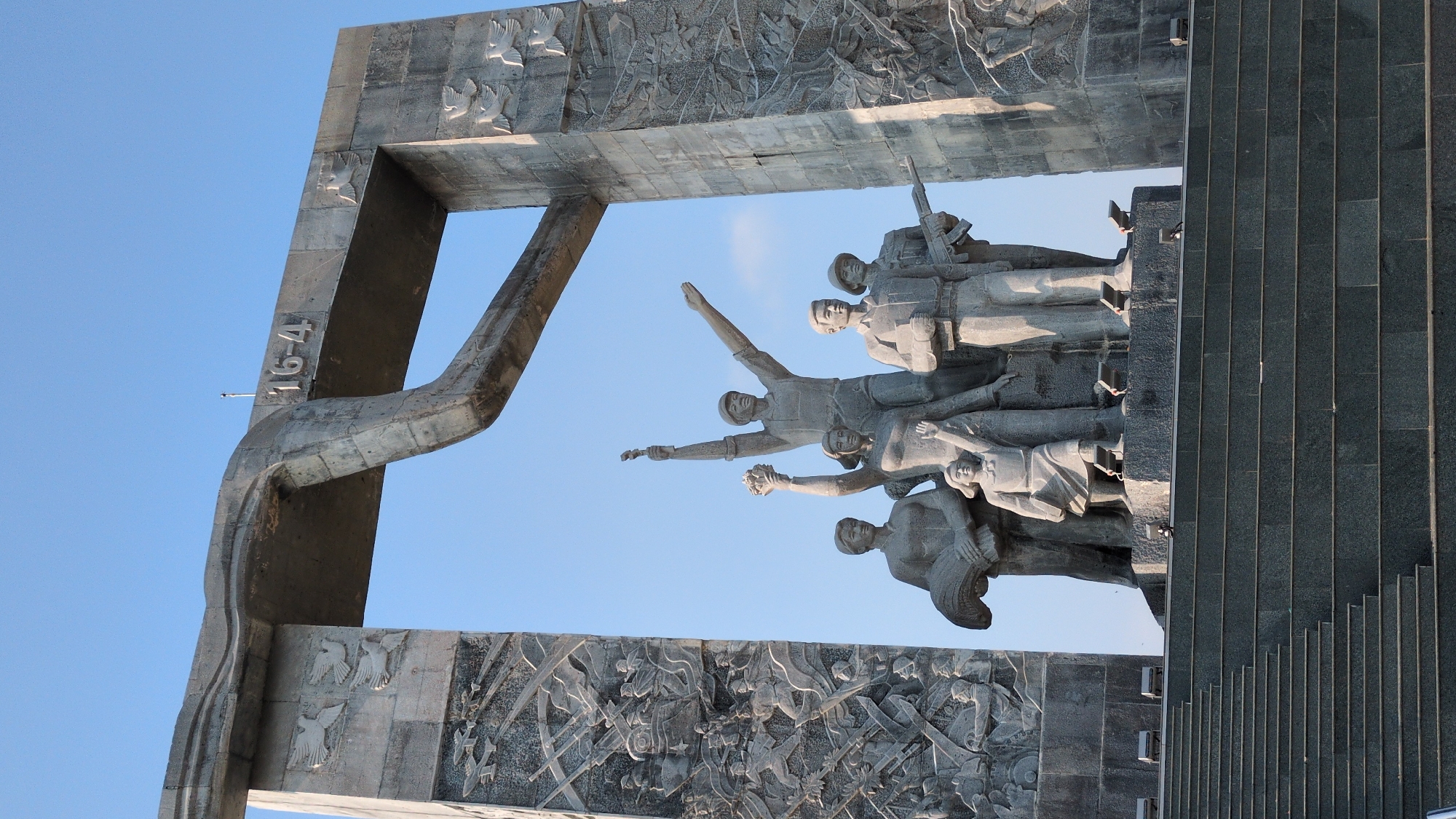 The image shows a concrete monument in Phan Rang–Tháp Chàm, Vietnam. The monument features a large archway with bas-relief carvings of battle scenes. Within the archway is a group of sculpted figures, possibly representing soldiers or civilians. To the right of the figures, there are gray stone steps. The sky visible through the arch is clear and blue. The date "16-4" is visible on a part of the archway.