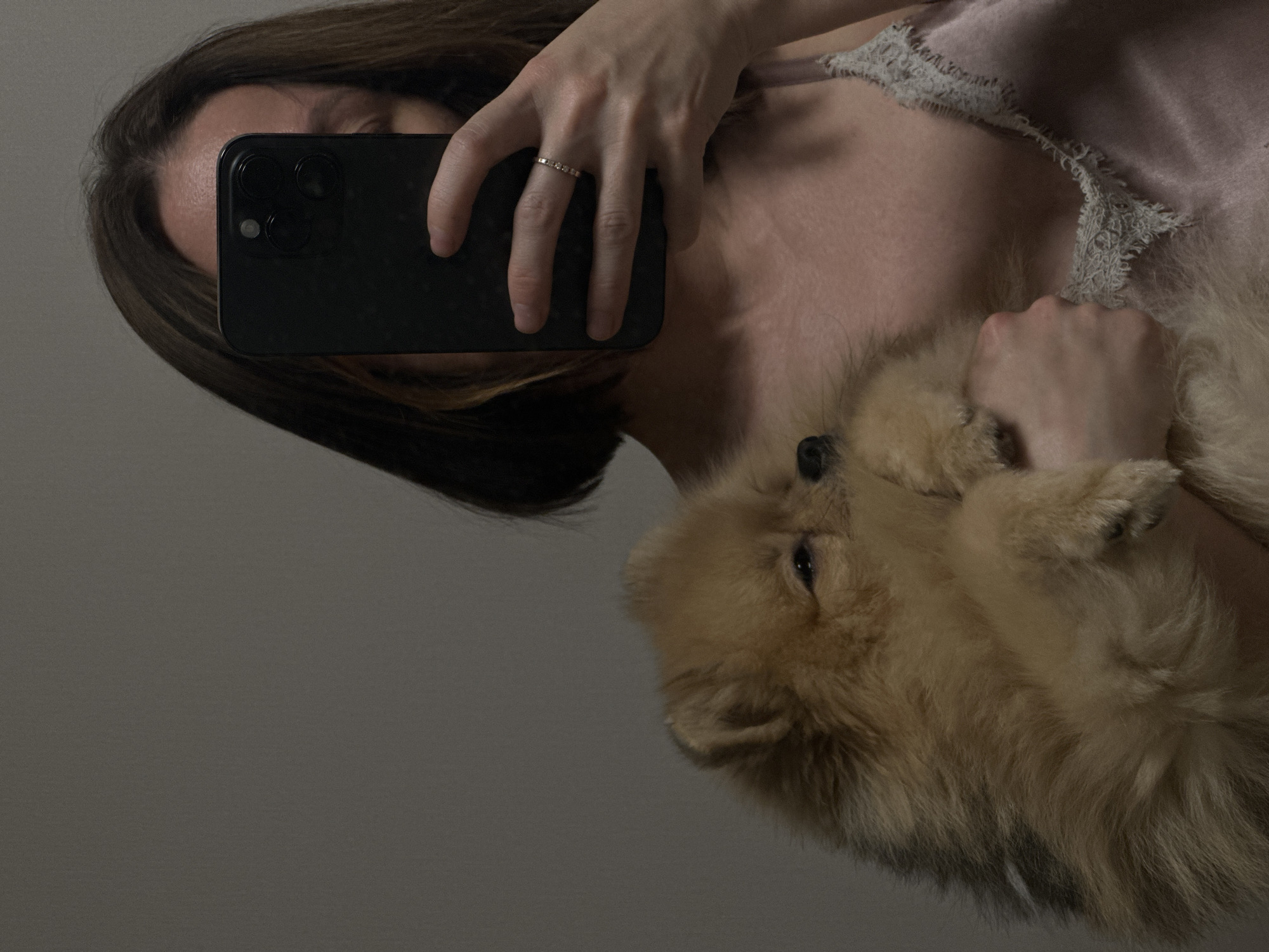 A woman is taking a selfie in a dimly lit room. She is holding a light brown Pomeranian dog in her arms. The dog is looking away from the camera, and the woman's face is partially obscured by her phone. She is wearing a pink satin camisole with lace trim. A silver ring is visible on her right hand. The background is a plain, neutral-colored wall. There are no discernible landmarks or features that would indicate a specific location within Votkinsk, Russia.