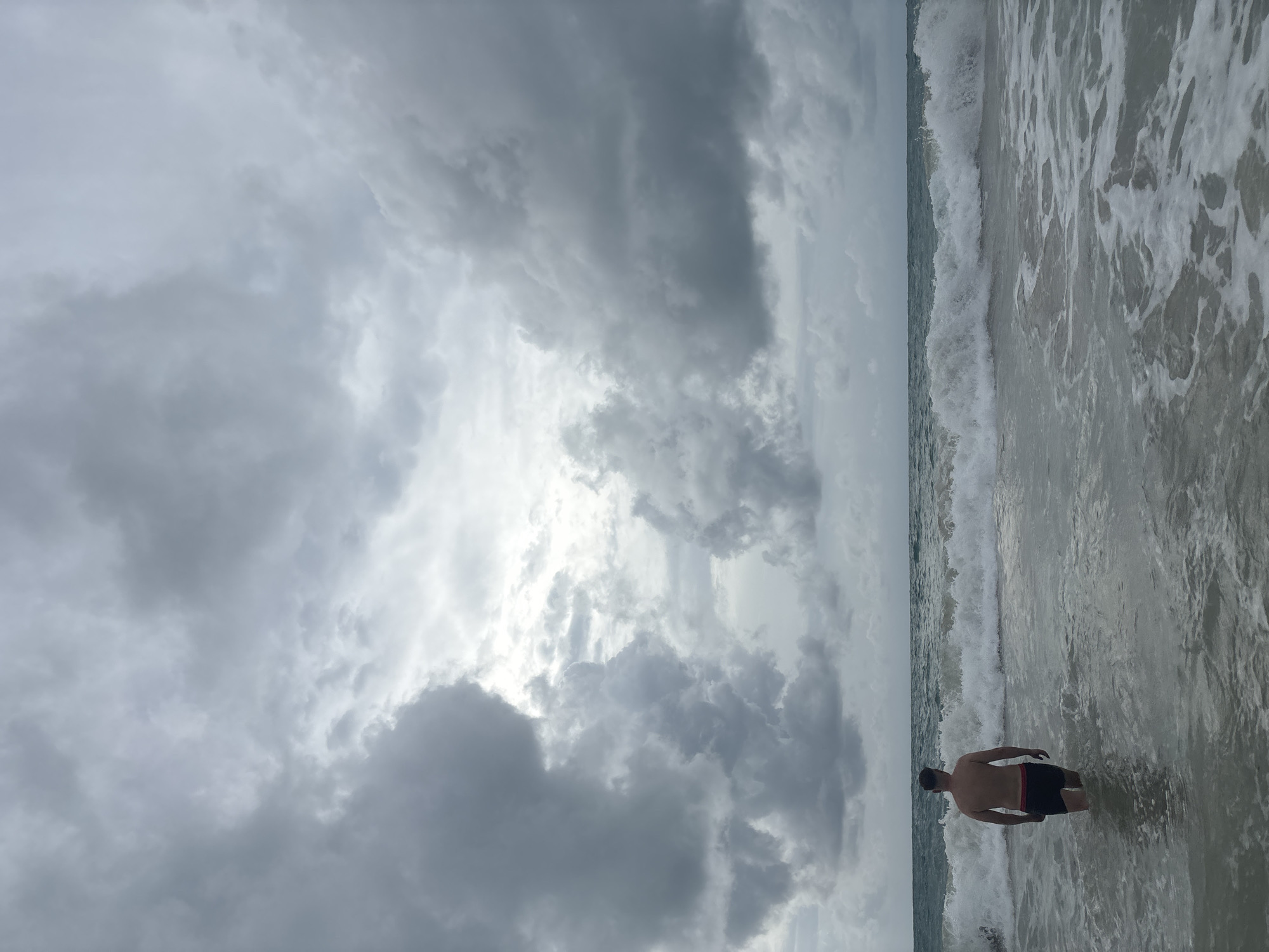A medium shot shows a person waist-deep in the surf at a beach. The location is Choeng Thale, Thailand, as indicated by the environment. The person is a male, facing away from the camera, wearing dark swim trunks. His skin tone is light. The sky is overcast, with heavy, gray storm clouds dominating the upper two-thirds of the image. The ocean is churning with white-capped waves breaking near the shore. The water appears murky, and the image is taken from an eye-level perspective. The overall mood is somber due to the stormy weather.