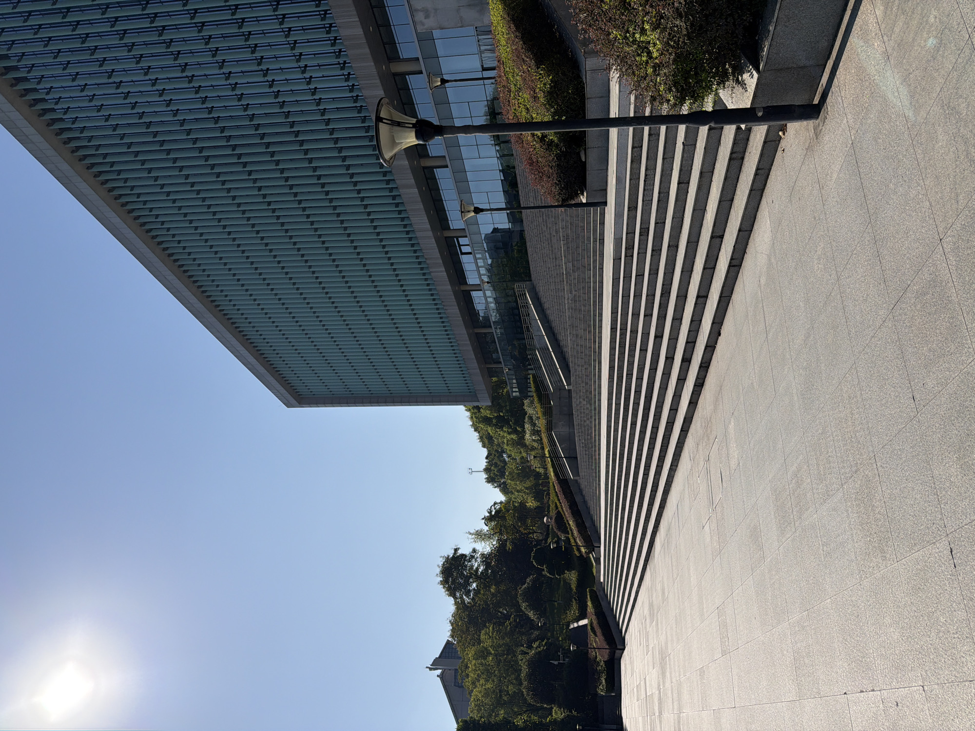 The image shows a bright, sunny day with a clear blue sky. In the foreground, a wide, tiled plaza leads to a long, wide staircase made of stone. The staircase ascends towards a modern building with a facade of vertical, greenish glass panels and a grey concrete frame. The building is large and imposing, with multiple floors visible.
To the left of the staircase and the building, there is a landscaped area with lush green trees and bushes. Several streetlights, with distinctive modern designs, are positioned along the path leading up the stairs. One streetlight is prominently featured in the mid-right of the image, casting a shadow on the pavement. The sun is visible in the upper left corner, creating a bright glare.
The scene appears to be an outdoor public space, likely a plaza or the entrance area of a public institution, university, or corporate building. The overall impression is one of modern architecture set within a well-maintained, green environment. The location of Jiangjin, China, is relevant to the architectural style and the landscaping which is typical of such regions. There are no people visible in the image.