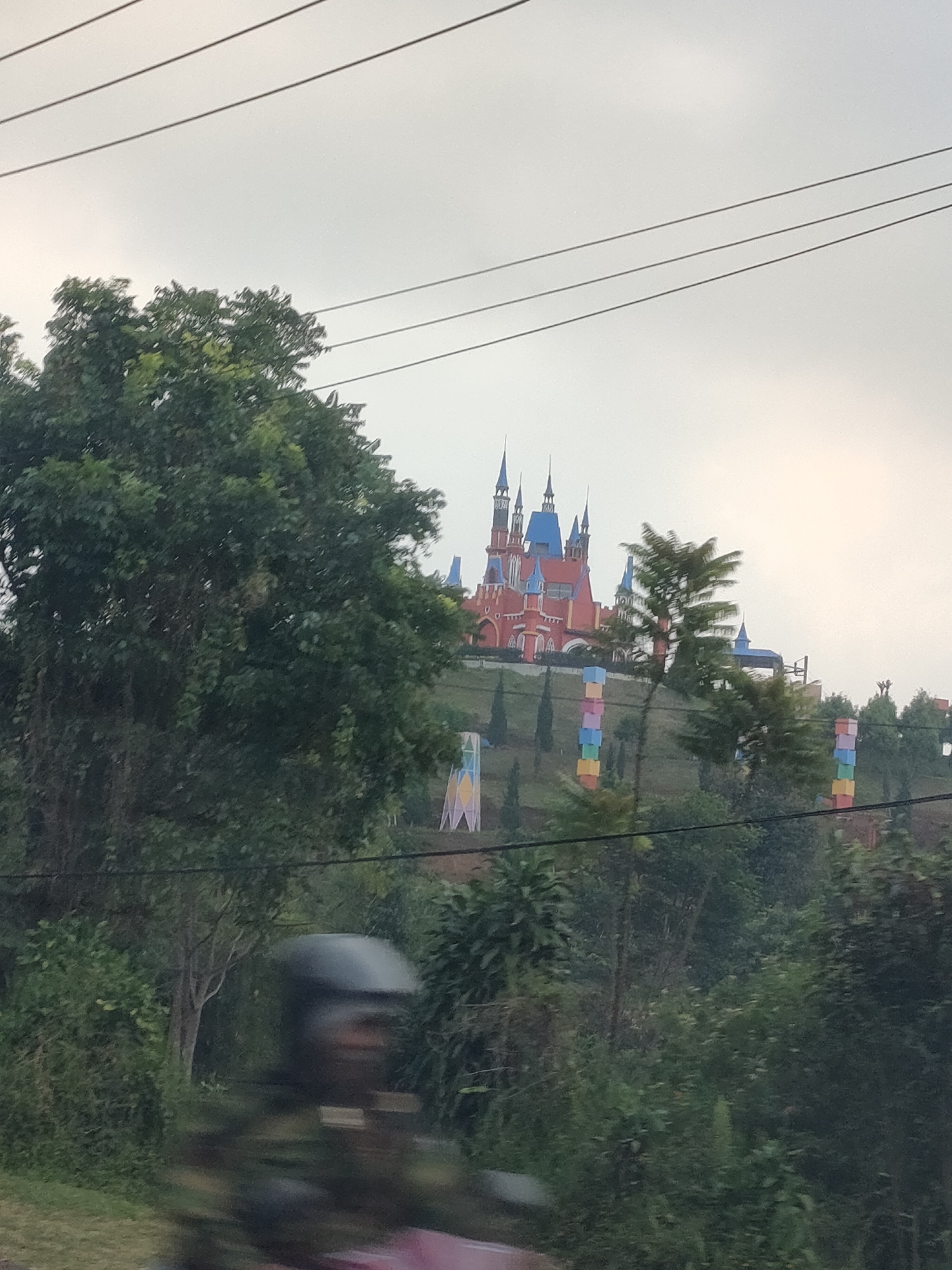 A colorful castle-like structure with blue roofs and red walls sits atop a hill in Subang, Indonesia. The castle is surrounded by lush greenery and several tall, thin poles with stacked colorful blocks. In the foreground, the blurry image of a person on a motorcycle wearing a black helmet is captured, suggesting the photo was taken from a moving vehicle. Overhead, power lines cut across the pale sky.