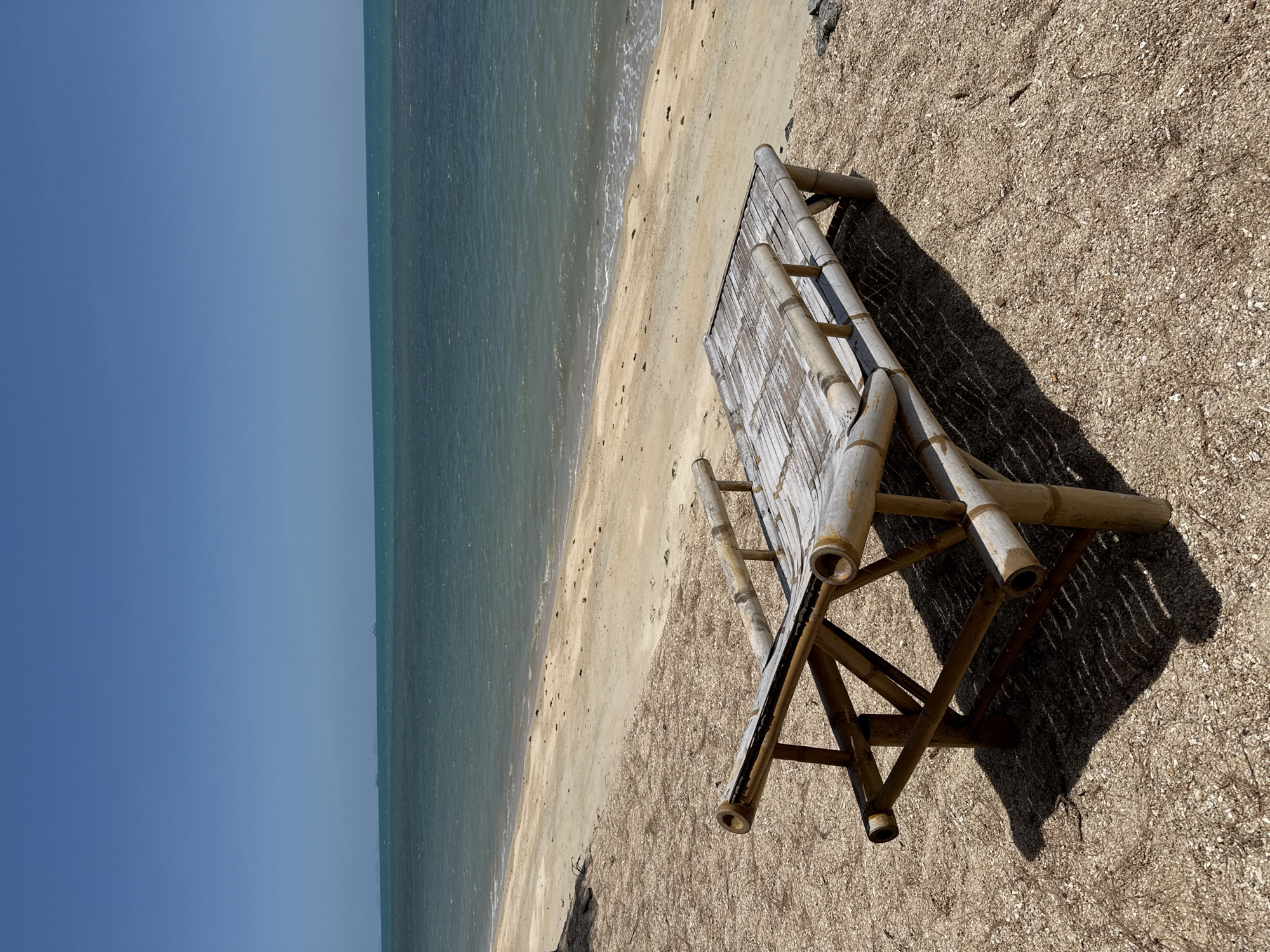 A lone bamboo lounge chair sits on a sandy beach in Ko Samui District, Thailand. The chair is made of natural bamboo poles and has a slatted surface. It is positioned facing the ocean, with the turquoise water stretching out to the horizon under a clear blue sky. The sand is light brown and speckled with small shells and debris. The chair casts a long shadow on the sand, indicating a bright, sunny day. The scene is peaceful and evokes a sense of relaxation and tropical paradise. There are no people or other objects of significance in the frame.