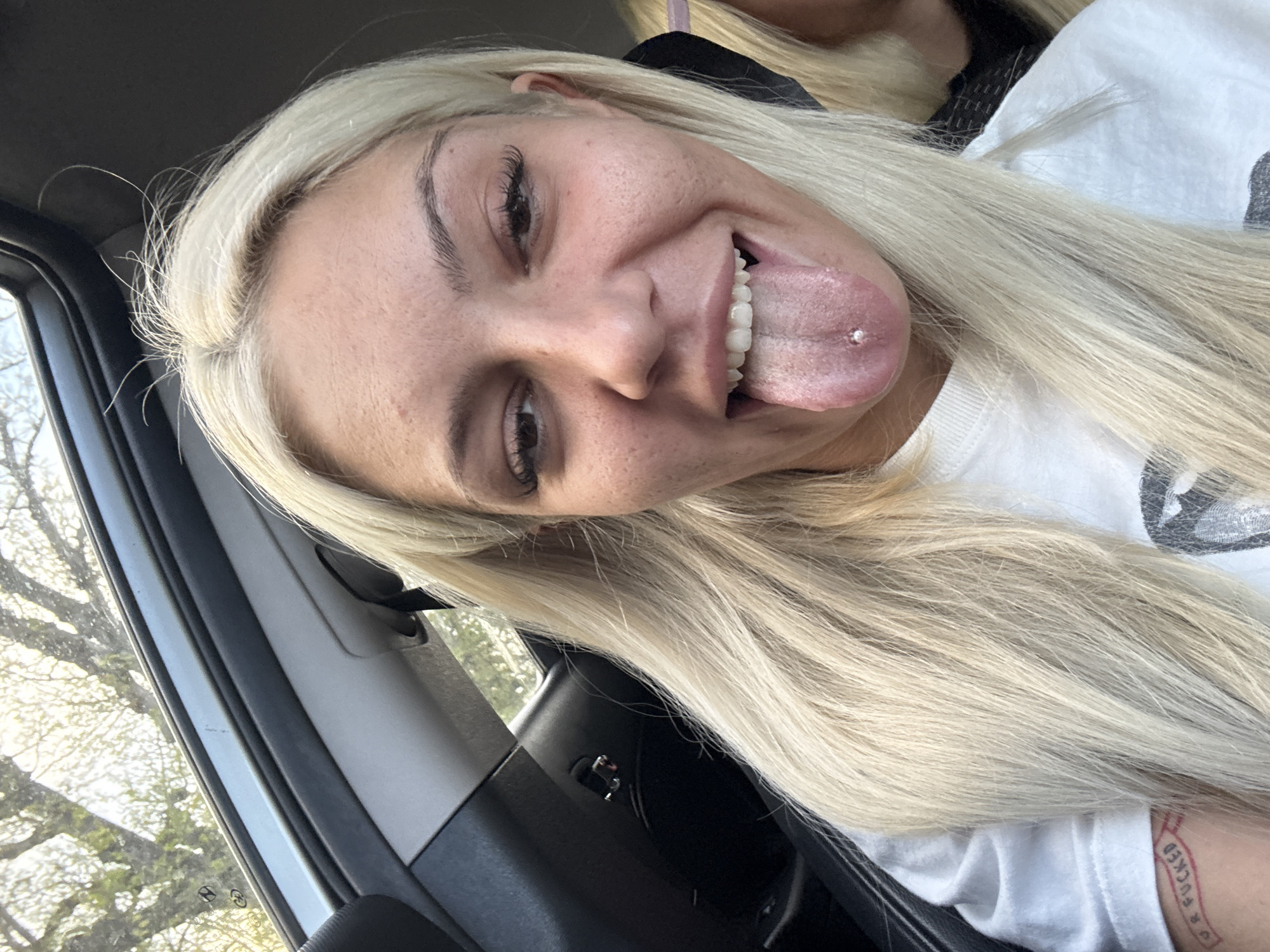 The image shows a young woman with blonde hair sticking out her tongue, which is pierced with a stud. She is wearing a white shirt. The setting appears to be inside a car. The exact location is not identifiable as Tulovo, Bulgaria.