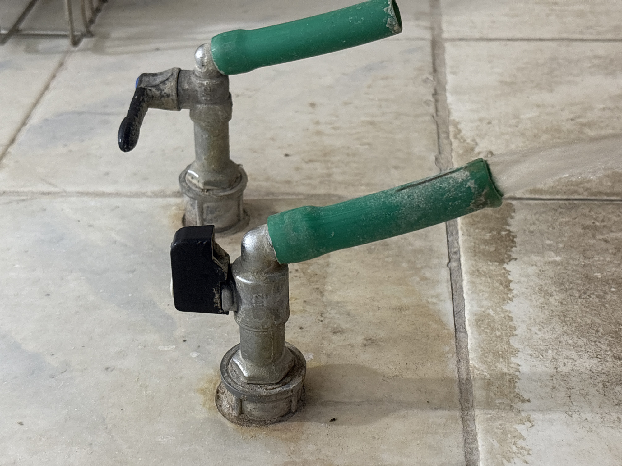 Two metal faucets are mounted on a tiled wall in Palimanan, Indonesia. Both faucets have green hoses attached to their spouts, and water is flowing from the faucet on the left. The wall is made of rectangular, light-colored tiles with visible grout lines and some staining, suggesting it is an outdoor or utility area. The faucet on the left has a black handle, while the faucet on the right has a black lever-style handle. The scene depicts a functional, albeit somewhat aged, water source.