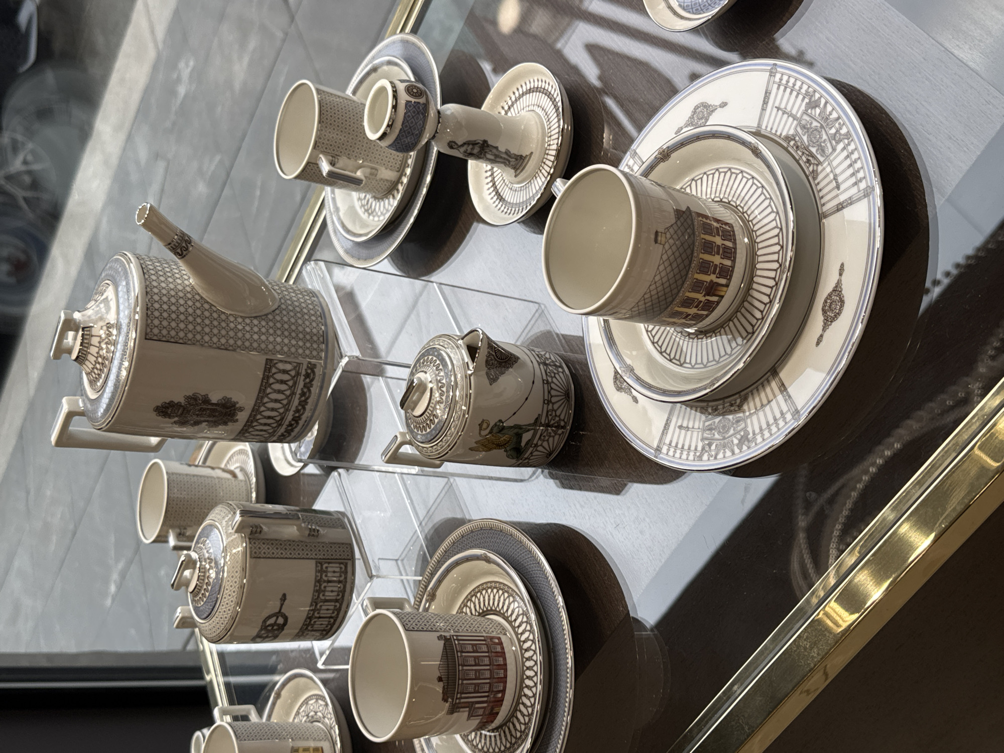 The image displays an array of fine porcelain tableware exhibited on a dark, reflective surface, presumably within a display case. The arrangement includes teapots, cups, saucers, plates, and sugar bowls, each adorned with intricate, classical patterns in white, blue, and gray. The objects are held in place by clear, acrylic stands. A bright, golden border is visible along the right edge of the display case. This high-end porcelain may be displayed in a shop or museum gift shop. The style of the pieces suggests it may be Russian Imperial Porcelain, such as from the city of Saint Petersburg.