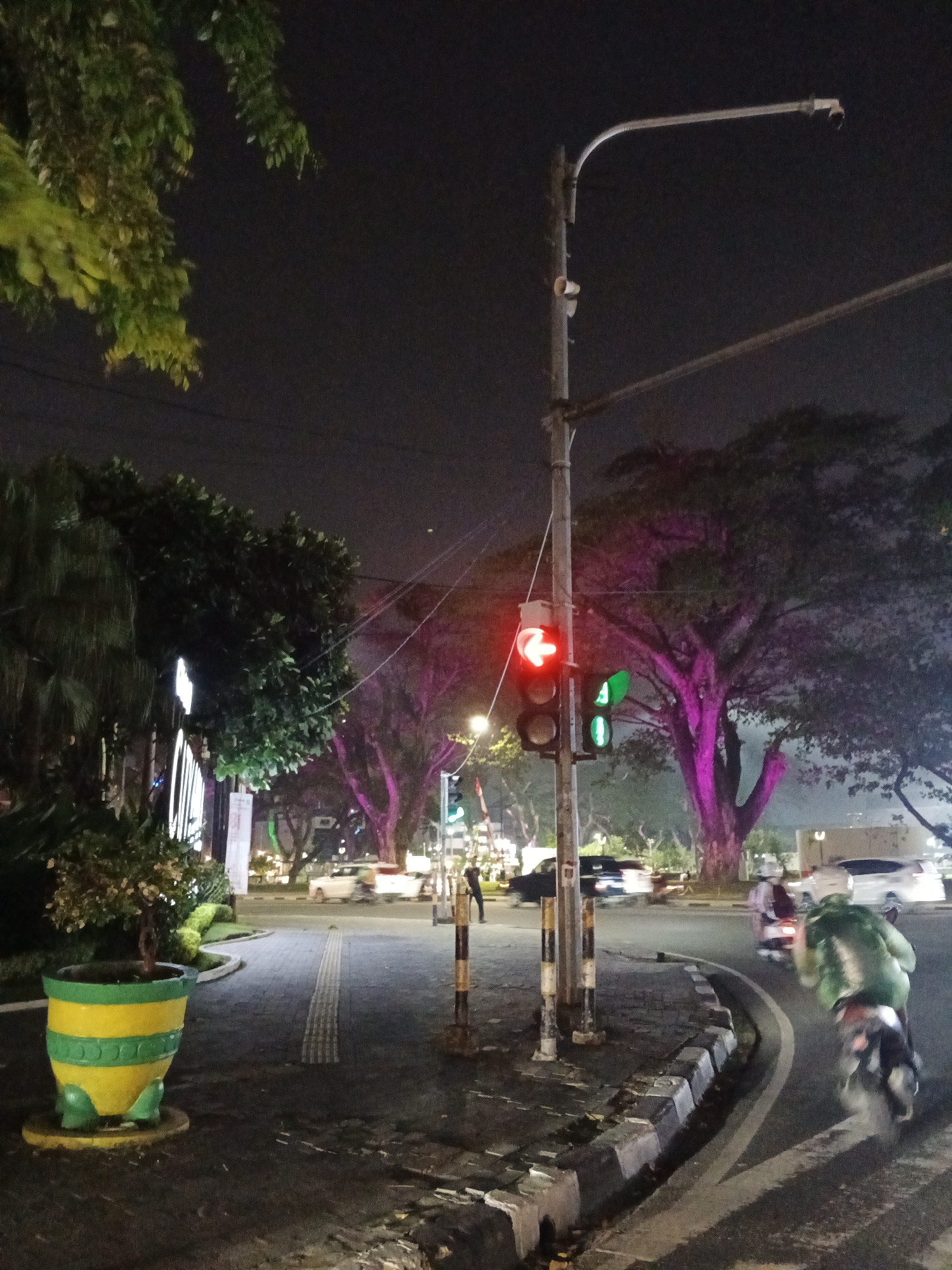 The image captures a night scene at a street intersection in the city of Medan, Indonesia. A traffic light is illuminated with a red arrow pointing left, indicating a restricted turn. Nearby, a green traffic light is also visible. A street lamp with a security camera attached extends over the intersection. Trees with foliage illuminated in pink are present in the background. On the right side of the road, a person is riding a motorcycle with a large green object strapped to their back, moving away from the camera. Several cars are visible in the middle ground, some appearing to be stopped at the intersection. In the foreground on the left, a decorative planter with yellow and green bands and frog-like feet holds a small plant. The ground is paved with interlocking bricks and concrete. The overall environment suggests an urban street at night.