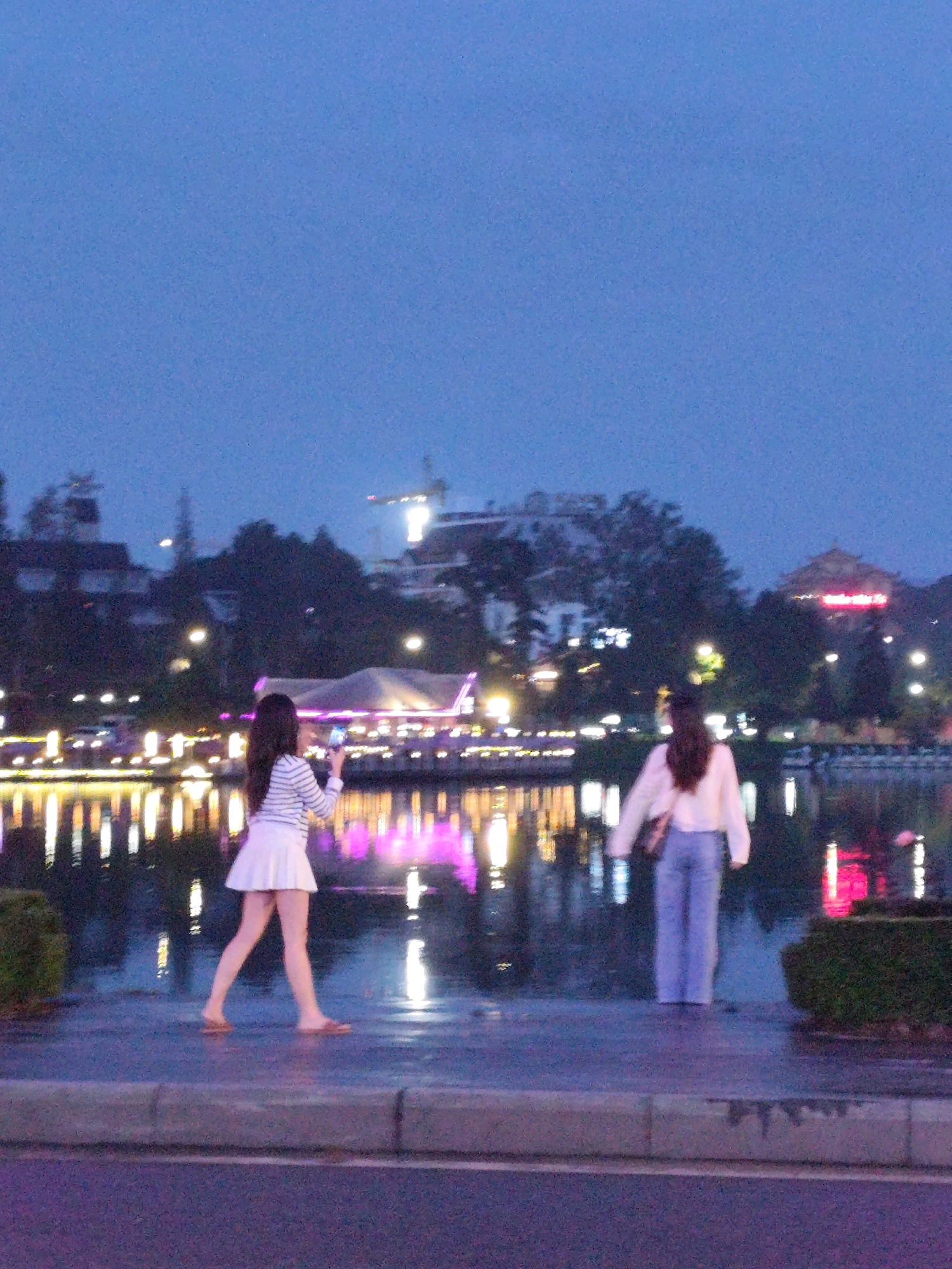 The image depicts Xuan Huong Lake in Da Lat, Vietnam, at dusk. Two young women are visible on the paved walkway adjacent to the lake. The woman on the left, facing away from the camera, is wearing a white pleated skirt and a horizontally striped long-sleeved shirt. She is holding a smartphone in her right hand, appearing to take a photo or video. The woman on the right, also facing away, is wearing light blue pants and a white long-sleeved top. She is carrying a dark shoulder bag. In the background, the lake reflects the vibrant lights of buildings and structures lining the shore, creating a shimmering effect on the water. The city skyline, with its buildings and trees, is dimly visible against the twilight sky, with a crane also present. The walkway and the surrounding area appear wet, suggesting recent rain.