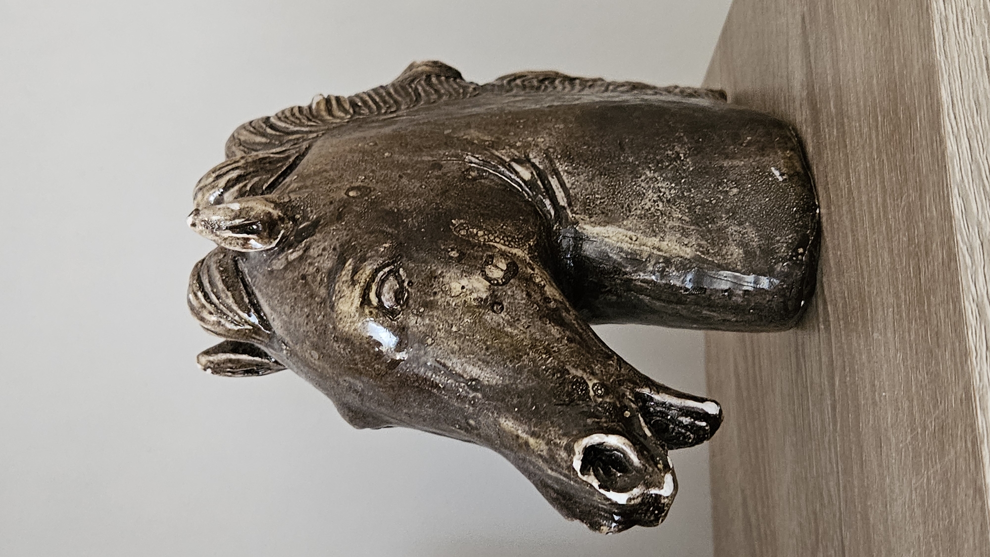 A close-up, side-profile photograph captures a dark, glossy ceramic horse head sculpture resting on a wooden shelf. The sculpture, with its open mouth and flared nostrils, exhibits a textured, rustic finish with lighter speckles. The horse's mane is depicted with carved wavy lines, and its eye is a dark, sunken socket. The background is a plain, light-colored wall, and the shelf beneath is made of light brown wood with visible grain. The lighting is even, highlighting the sheen of the ceramic. The location of this object is not determinable from the image.
