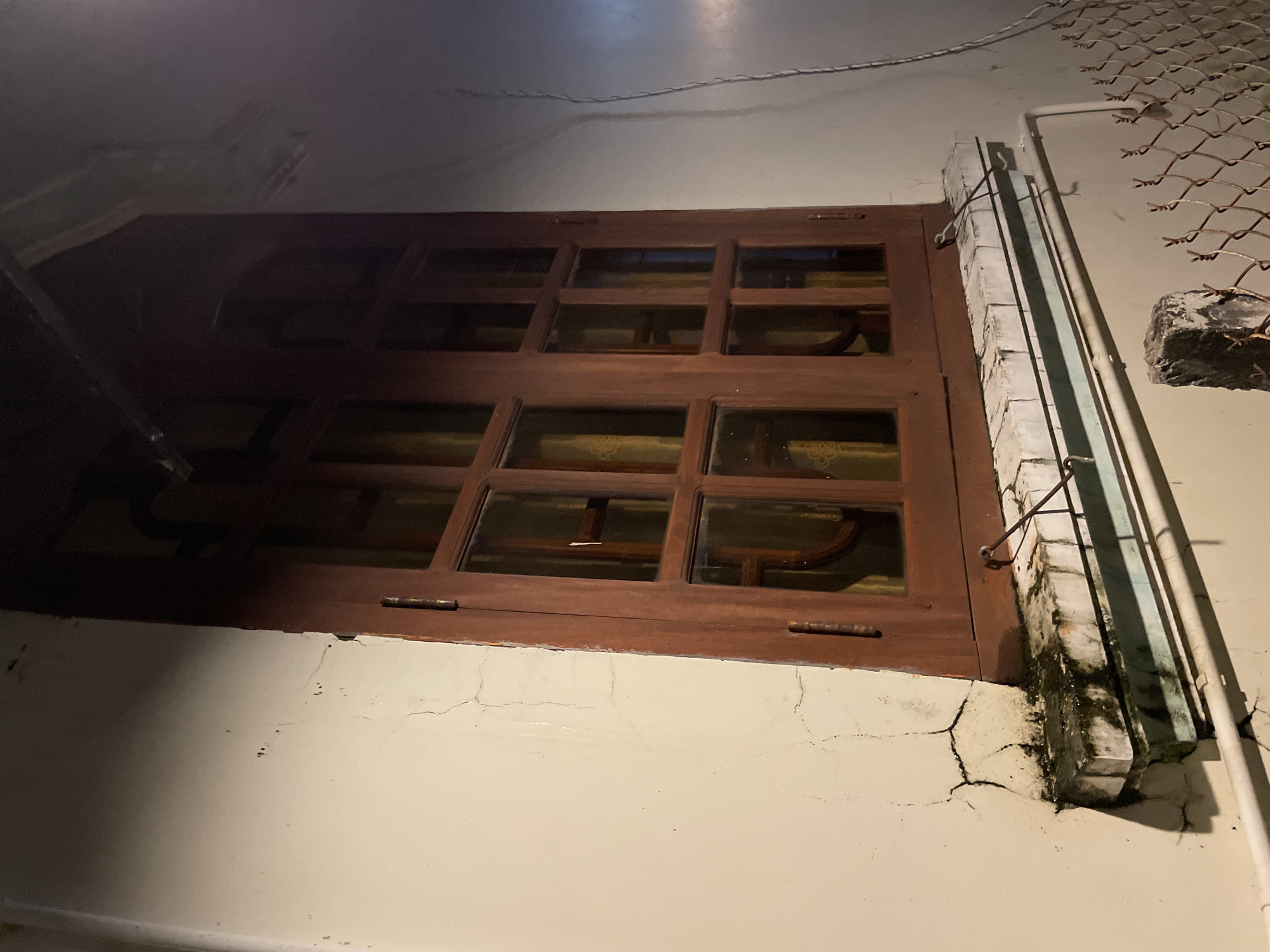 A wooden-framed window with multiple panes is installed on a light-colored, cracked exterior wall. The window has dark brown panes and is set above a stone windowsill that shows signs of wear and mildew. A chain-link fence runs along the bottom of the frame, with a concrete post supporting it. The scene appears to be outdoors, potentially at night given the lighting and shadows. There is no indication of the specific location within Phu Thuy Ward, Vietnam, beyond the general impression of a building exterior.