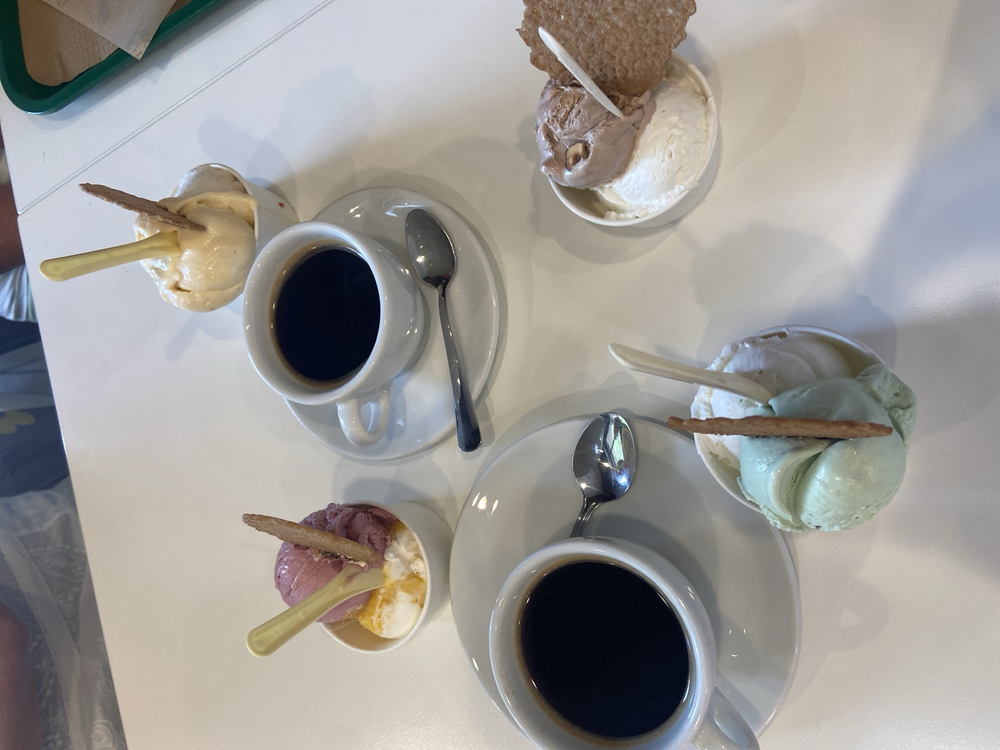A high-angle shot captures a tabletop scene featuring four small bowls of gelato and two cups of black coffee, along with spoons and thin, rectangular wafers. The gelato bowls hold different flavor combinations. From the top left going clockwise, there is a bowl with what appears to be light yellow gelato, then a bowl with chocolate and vanilla gelato topped with a large, round wafer, a bowl containing green and white gelato, and lastly a bowl with purple and yellow gelato. All the bowls are paired with a spoon. Two coffee cups, each on a saucer, are placed near the bowls of gelato. The scene takes place on a light colored table, but there is not enough context to determine the location in Lecco, Italy.