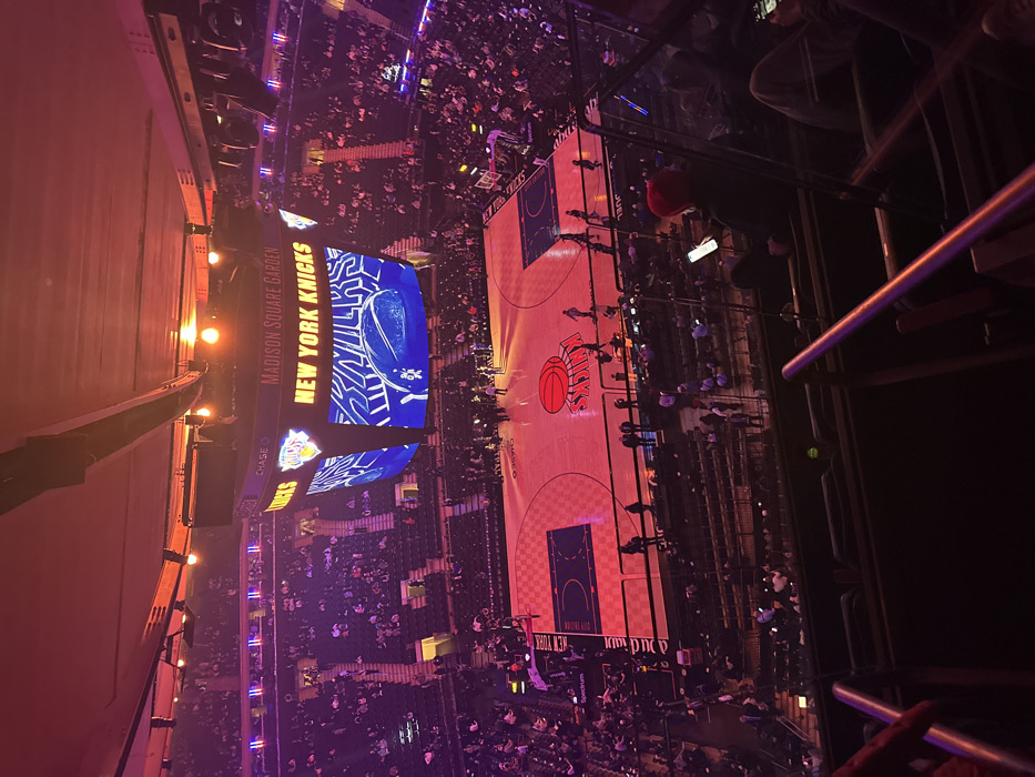 An elevated view of Madison Square Garden showcases a basketball court under bright lights. The court prominently features the New York Knicks logo. A large screen displays "New York Knicks" above the court, while stadium seating is filled with spectators. Metal railings are visible in the foreground. The image appears to be taken from a high vantage point within the arena.