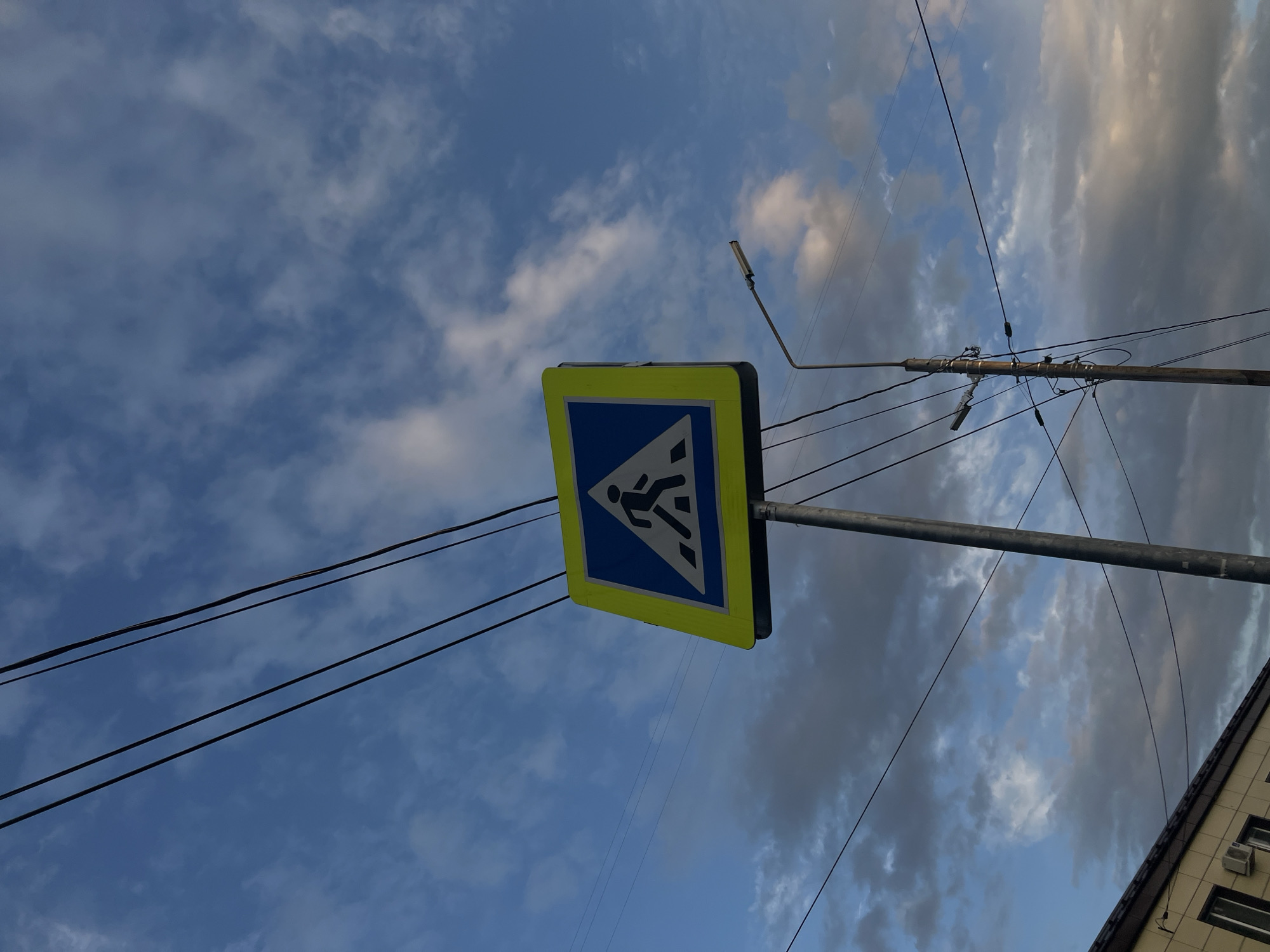 A pedestrian crossing sign is depicted against a cloudy blue sky. The sign is square with a yellow border and a blue background, featuring a black silhouette of a person walking. The sign is mounted on a metal pole. Several power lines crisscross the sky, and a lamppost is visible to the right of the sign. In the lower left corner, the corner of a building with a beige facade and dark brown trim is visible. The sky has a mix of white and gray clouds, with some areas of blue sky visible. The image appears to be taken from a low angle, looking up at the sky and the sign. The general context of the scene is urban, with the presence of the building, street sign, and power lines suggesting a street or road in Ulyanovsk, Russia.