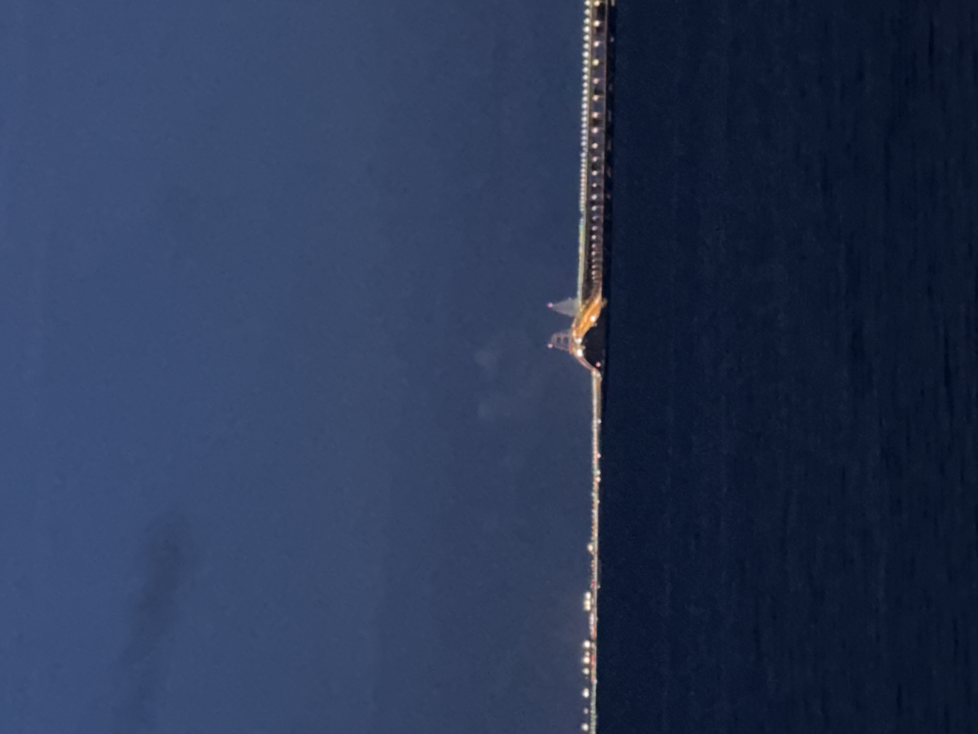 The image depicts a long, illuminated bridge spanning a body of water at dusk. The bridge is lit by numerous lights along its length, creating a bright ribbon against the dark, twilight sky and the deep blue water. In the distance, on the horizon, faint lights suggest a city or coastal area. The sky is a deep, dark blue with some faint cloud formations visible. The water is calm with gentle ripples. The bridge appears to be a significant infrastructure project, likely a road or railway bridge, with distinctive support structures or towers visible towards the center. The overall impression is one of a vast, man-made structure integrated into a natural landscape at a transitional time of day.