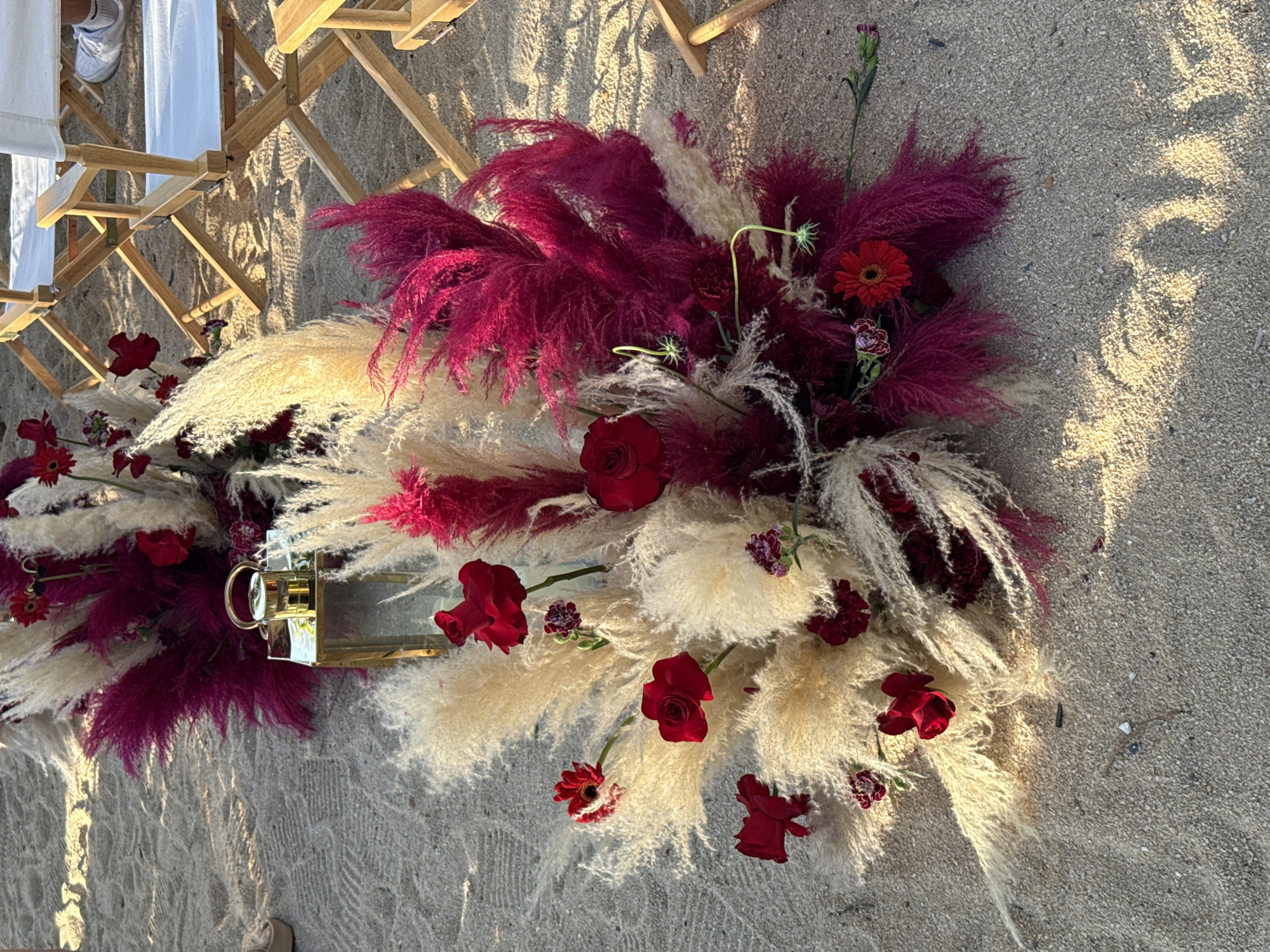 A floral arrangement of pampas grass and dark red roses is set on a sandy beach in Baan Bo Phut, Thailand, with folded wooden chairs in the background. The arrangement features fluffy, cream-colored pampas grass interspersed with vibrant, fuchsia-colored plumes and dark red roses. A small, golden lantern with a glass enclosure sits on the sand amidst the flowers. The sand is textured and shows patterns of light and shadow from the sunlight. In the background, two white canvas director's chairs with wooden frames are visible, and the legs of a person wearing white sneakers are in the lower right corner. The scene appears to be outdoors, likely on a beach, given the sand and the casual seating.