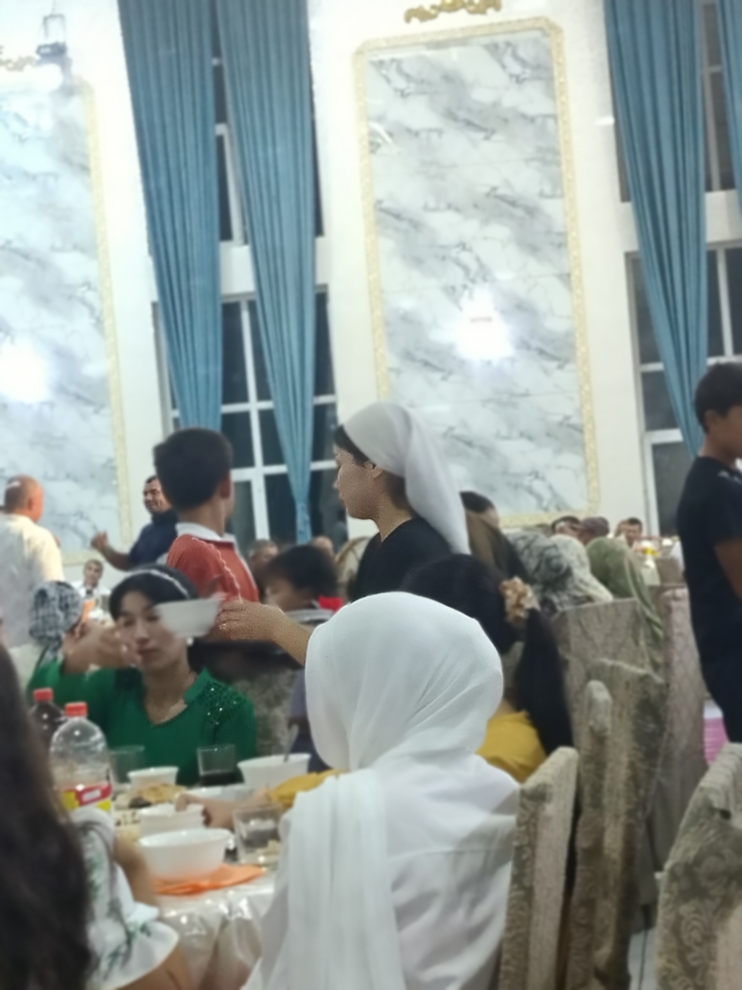 A medium shot captures a group of people seated around tables in what appears to be an indoor event space. The lighting is somewhat dim, with ambient light illuminating the scene. In the foreground, a person wearing a white headscarf is mostly out of frame, their back to the camera. To their left, a young woman in a green top is looking down at a bowl on the table, her right hand raised as if holding something. A bottle of liquid with a red cap is visible on the table near her. Further back, a young boy wearing an orange shirt is turned away from the camera, looking towards the left. Behind him, a man in a dark shirt is visible, giving a thumbs-up gesture. The background features tall, blue drapes and decorative wall panels with a marble-like pattern. Several other individuals are seated at tables, some partially obscured. The overall impression is of a gathering or meal taking place in a formal setting.