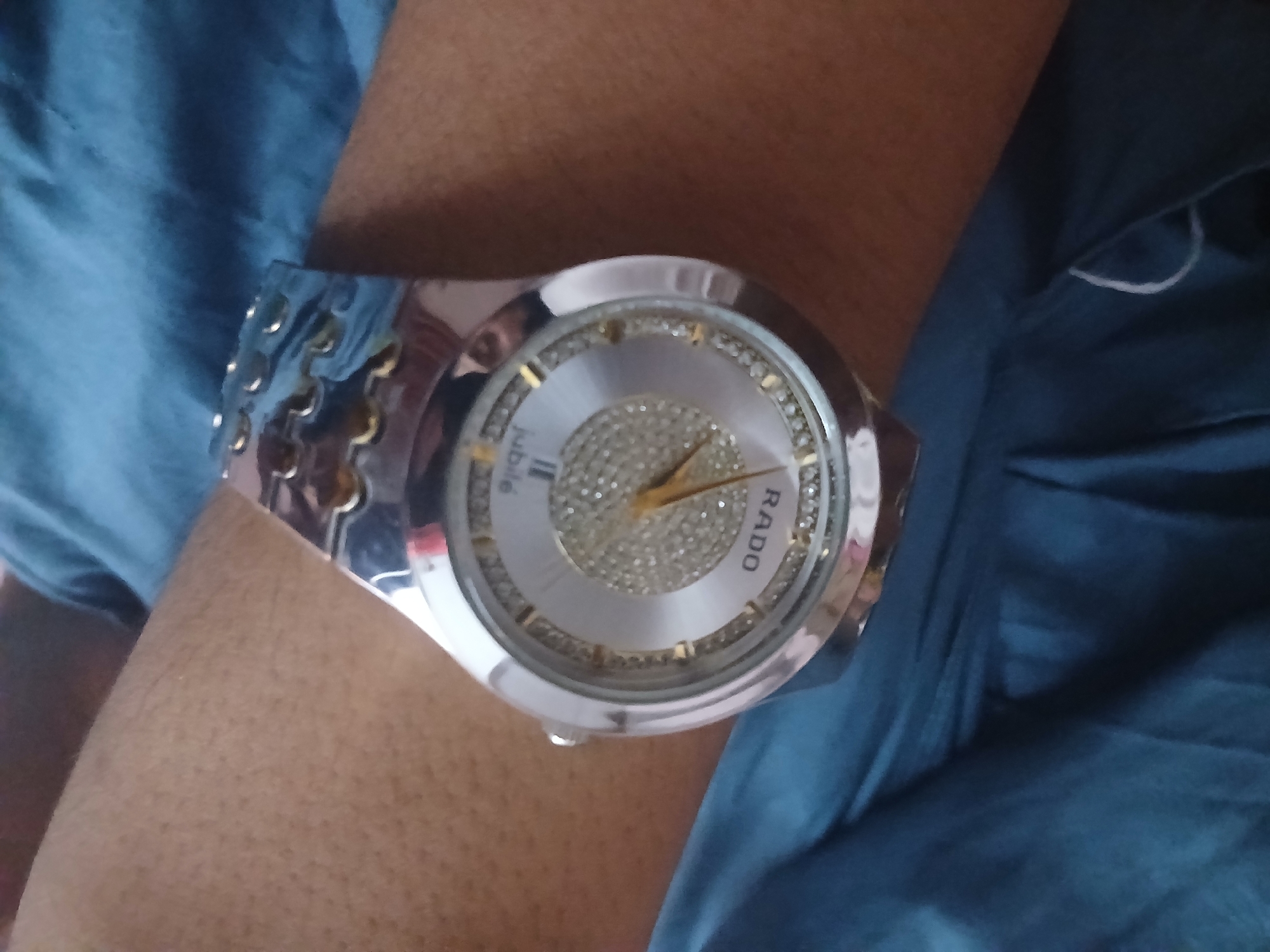 An overhead shot captures a Rado Jubilee watch on a person's wrist, adorned with diamonds. The watch is silver, with a silver face and a shimmering diamond-studded inner circle. The hands of the watch are gold. The person's wrist, a light brown color with visible hair follicles, is positioned against a dark blue, textured fabric, possibly clothing or a surface. The background is blurred, suggesting a close-up perspective focused on the watch. The image's lighting is somewhat uneven, with the watch face being brightly illuminated and some areas of the fabric appearing darker. No specific location in Peshawar City Tehsil, Pakistan is identifiable from the image.