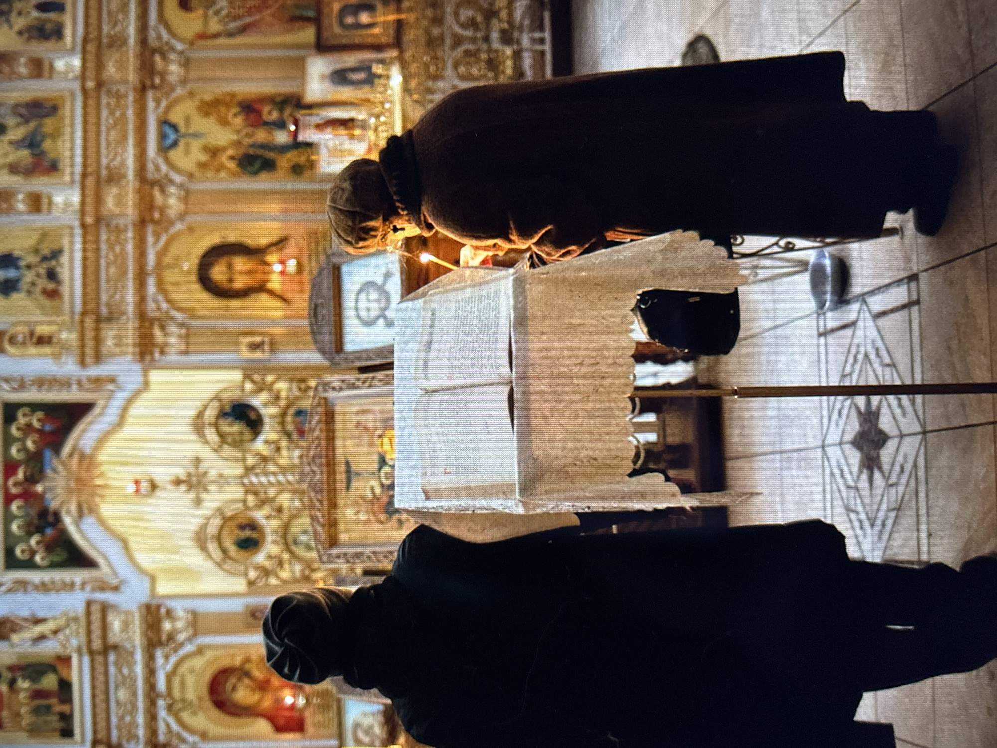 Two individuals, facing away from the camera, stand in an Orthodox church in Pastavy, Belarus. The person on the left wears a dark hooded garment and a dark beanie. The person on the right wears a dark coat and a brown knit hat. Both are positioned behind a stand displaying an open book, adorned with a lace cloth. A lit candle is visible near the person on the right. The background features religious icons and ornate architectural details typical of an Orthodox church interior. The floor is tiled with a geometric pattern.