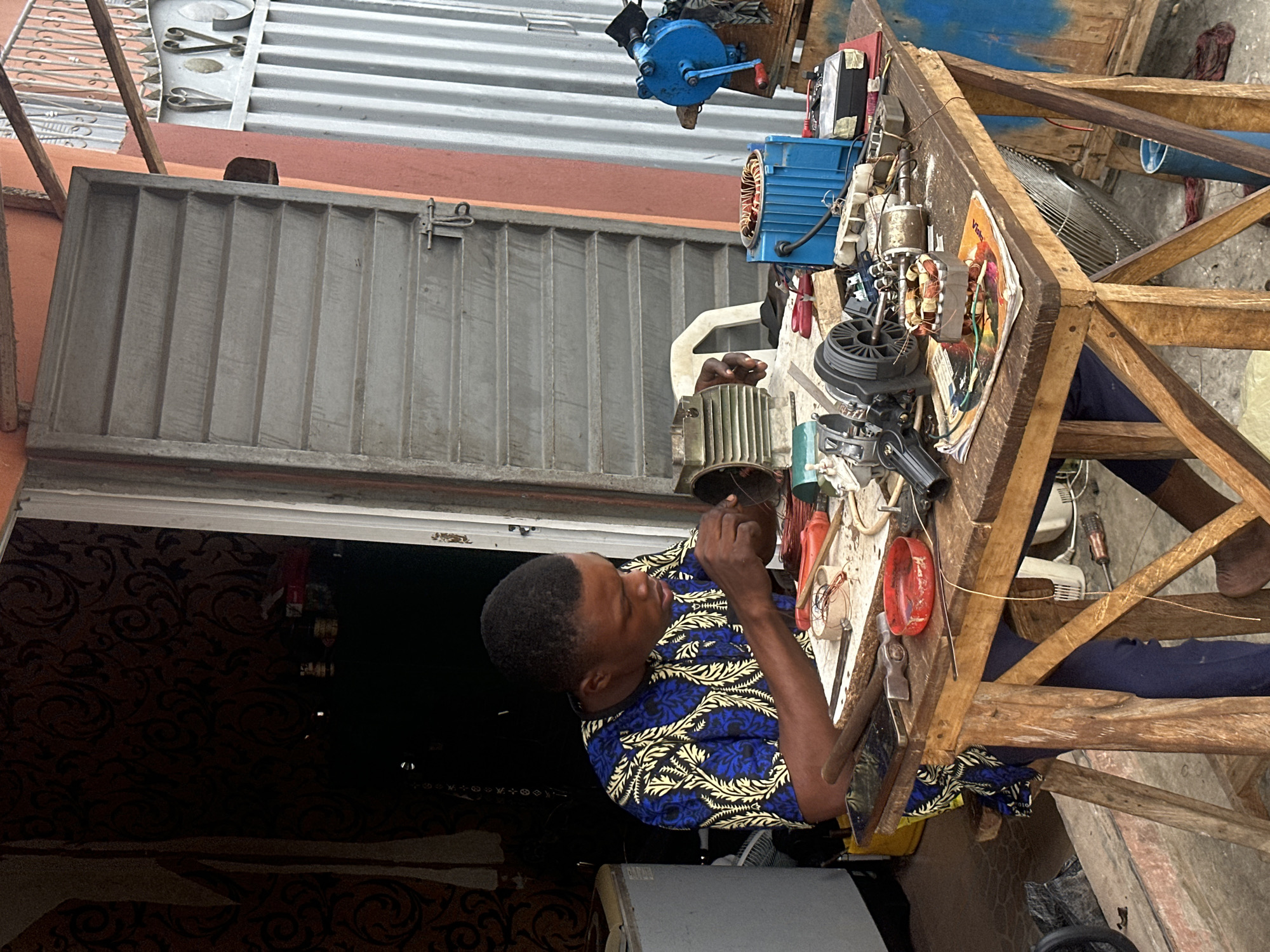 In Badagry, Nigeria, a young man is working on an electrical motor at a workbench outside a shop. He is wearing a blue and yellow patterned shirt and dark pants. His hands are engaged in the intricate work of repairing the motor, with various tools and components spread across the wooden table. These include screwdrivers, wires, tape, and dismantled parts of electrical devices. The environment appears to be an open-air workshop, with a corrugated metal roof structure and a metal door partially open, revealing a dark interior with patterned wallpaper. The scene suggests a skilled tradesman diligently performing his work in a typical Nigerian urban setting.