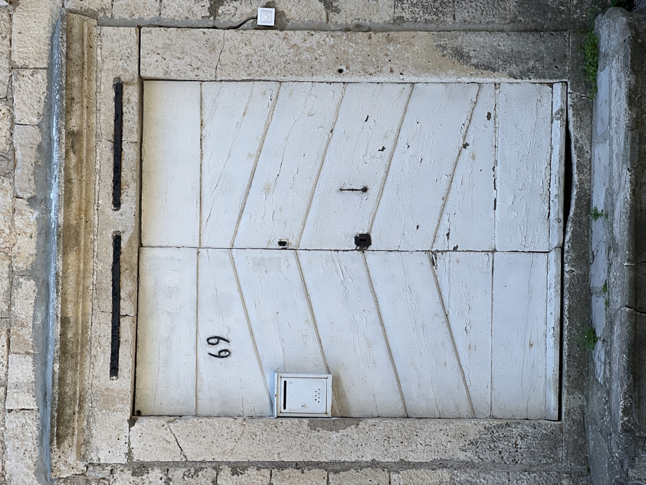 The image presents a static, head-on view of an aged, white double-leaf wooden door, designated by the house number "69", situated within a weathered stone architectural frame in Bogdašići, Montenegro. The door's white paint is extensively chipped and peeled, revealing the underlying material and indicating significant exposure. Each door leaf is composed of multiple horizontal planks, arranged to create a chevron or diagonal pattern across the central sections. A small, white, rectangular metal mailbox with lower perforations is affixed to the upper left door leaf, positioned below the "69". The right door leaf features a dark, prominent latch or lock mechanism, with a corresponding dark pull handle on the left leaf. Two recessed black horizontal metal bars are set into the stone lintel directly above the door. The surrounding wall is constructed from irregularly shaped, light-colored stone blocks. To the right of the door frame, a small, white rectangular electronic device, likely a doorbell or intercom component, is mounted. A worn stone step or threshold forms the base of the entrance, with sparse green vegetation emerging from cracks in the stone. The scene is illuminated by direct sunlight, highlighting textures and casting subtle shadows.