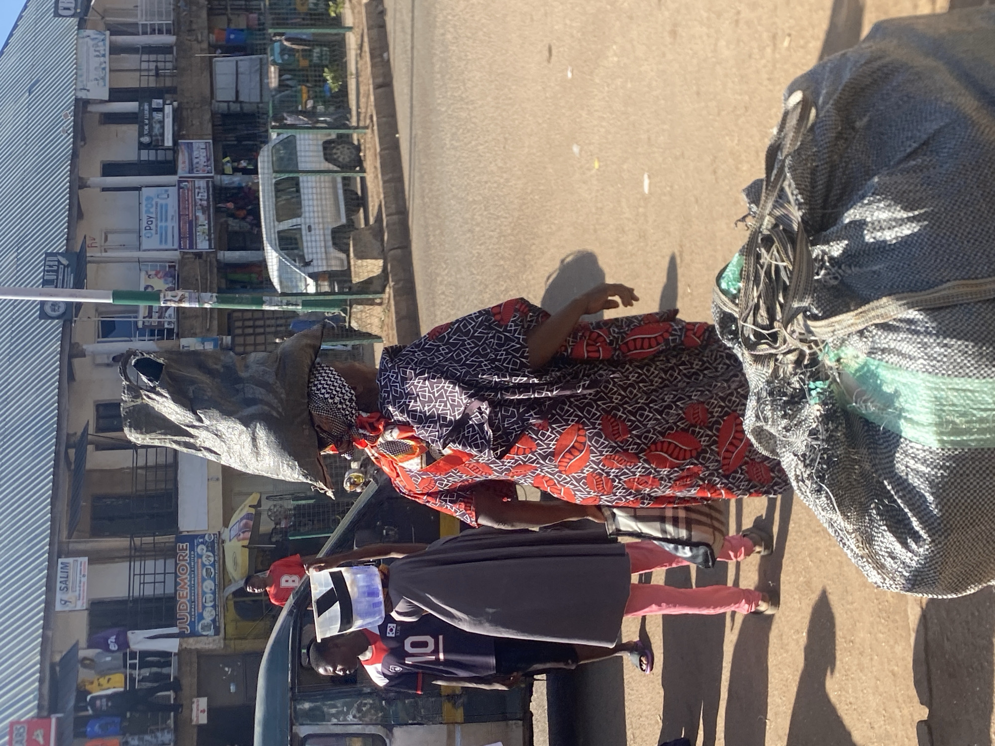 The image captures a street scene in Bukuru, Nigeria, with a focus on pedestrians and the surrounding urban environment. In the foreground, a woman is seen from behind, carrying a large, dark-colored sack balanced on her head. She is wearing a vibrantly patterned red and black dress with a white and black headscarf. To her left, a person in pink trousers and a grey covering is carrying a white plastic container on their head. A man in a dark blue jersey with the number "10" on the back stands nearby. In the background, several buildings with corrugated metal roofs line the street, displaying various signs, including "SALIM," "JUDEMORE," and "PayPOS." A white van is parked on the right side of the road, and other vehicles and people are visible further down the street. The ground is paved with asphalt, and sunlight casts shadows, indicating a bright, possibly midday, lighting condition. The overall scene depicts a bustling marketplace or thoroughfare.