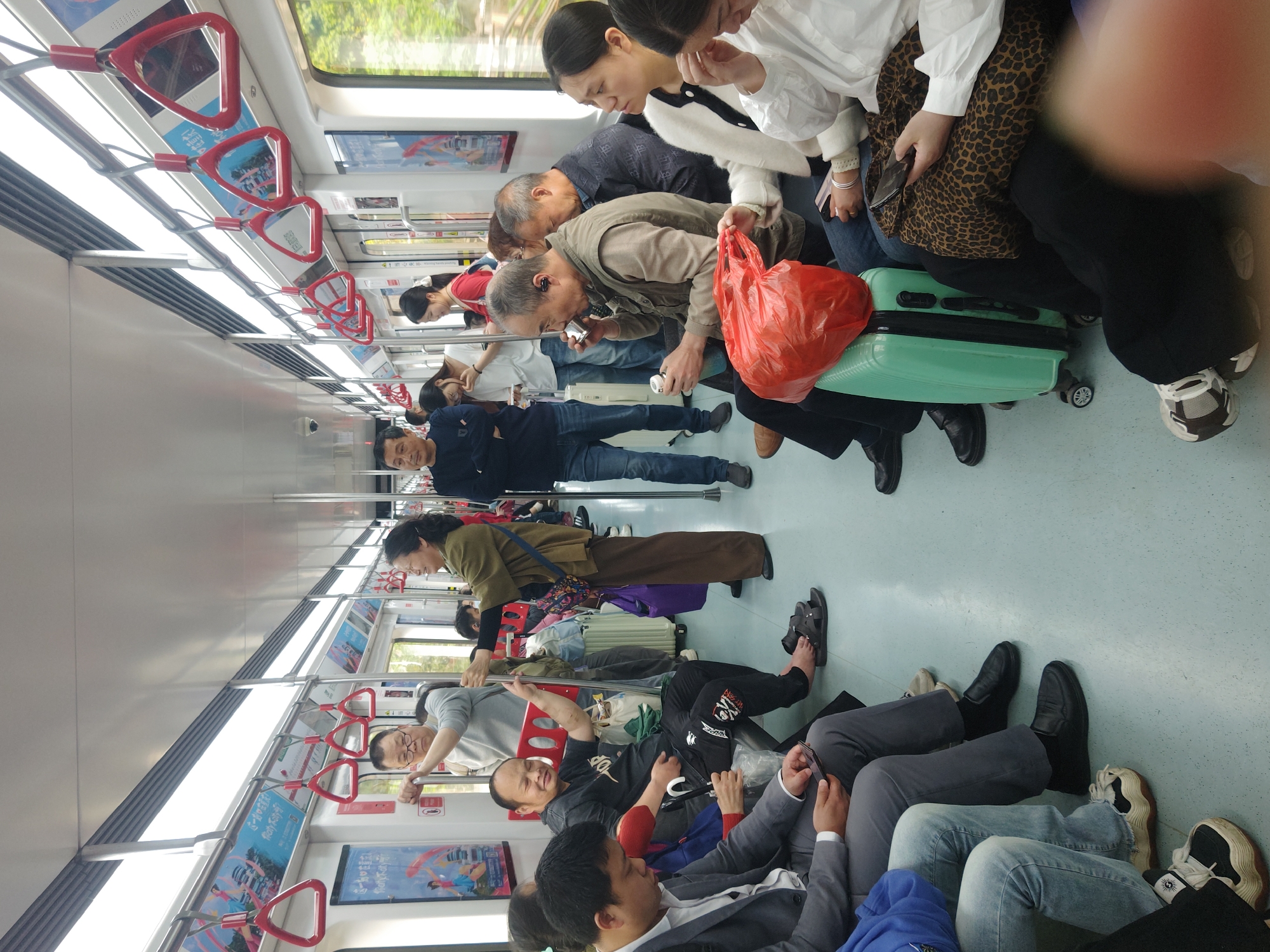The image shows the interior of a crowded subway car. Several passengers are seated or standing, with some holding onto overhead straps. There is luggage, including a light green suitcase and a red plastic bag, in the aisle. People are dressed in various casual and business attire. Some are looking at their phones. The scene is well-lit, and the subway car appears to be in motion, with a blurry view visible through the windows.
