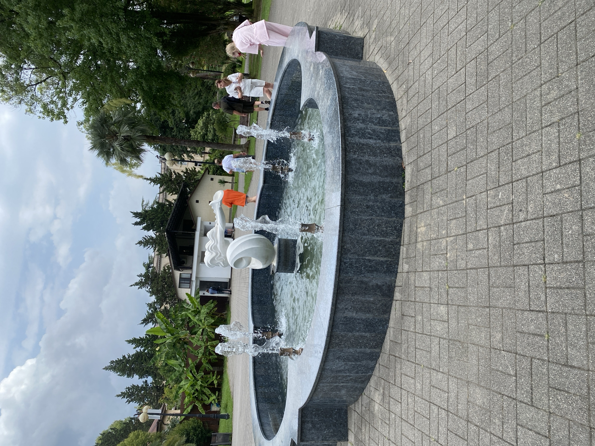 The image shows a fountain in a park in Sokhumi, Abkhazia. The fountain is made of dark gray stone and has a sculptural element in the center. Water is spraying from several spouts, creating a lively atmosphere. Several people are visible around the fountain, some walking and some standing and talking. The background features lush green trees, a building, and a cloudy sky. The ground around the fountain is paved with gray interlocking bricks.