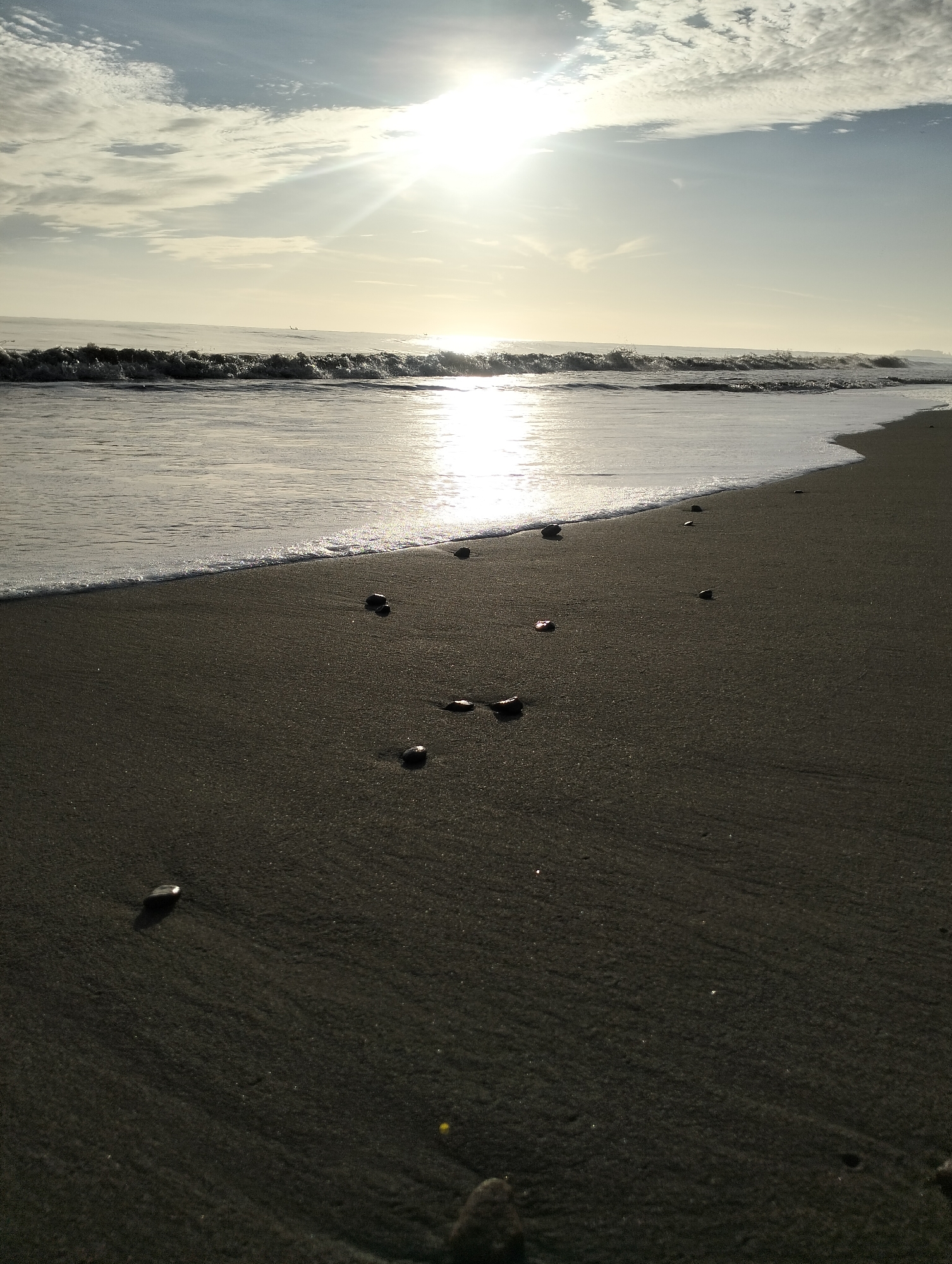 The photograph captures a beach scene, possibly in Sagoe, Indonesia, at dawn or dusk. The sun shines brightly through a thin layer of clouds in the sky, creating a strong reflection on the water. The beach has dark sand and scattered pebbles. Gentle waves meet the shore, leaving a trail of white foam. There are no people visible in the frame.