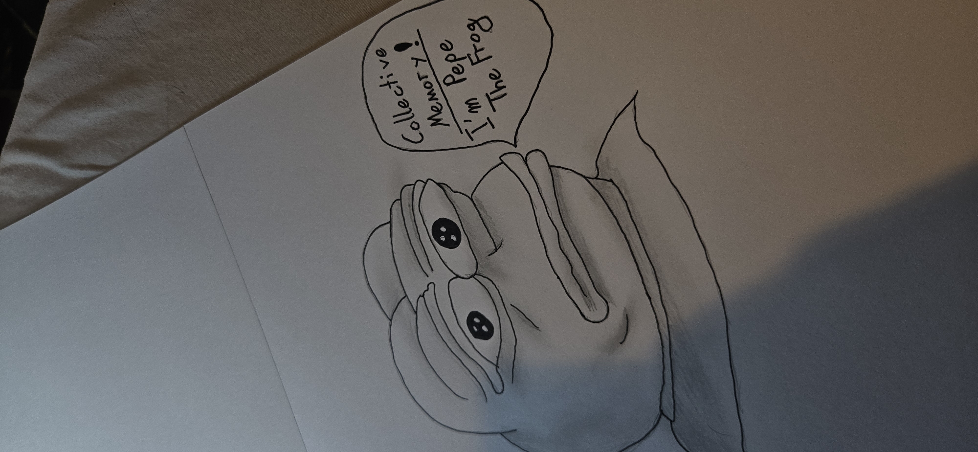 A pencil drawing depicts Pepe the Frog, an internet meme character, with a pensive expression. The frog is rendered in black and white, with shading to give it depth. To the right of the frog, a speech bubble contains handwritten text that reads: "Collective Memory! I'm Pepe The Frog". The drawing is on a sheet of white paper, which is placed on a textured, light-colored fabric. The background suggests the paper is part of a larger drawing or notebook. The overall composition is from a slightly elevated angle.