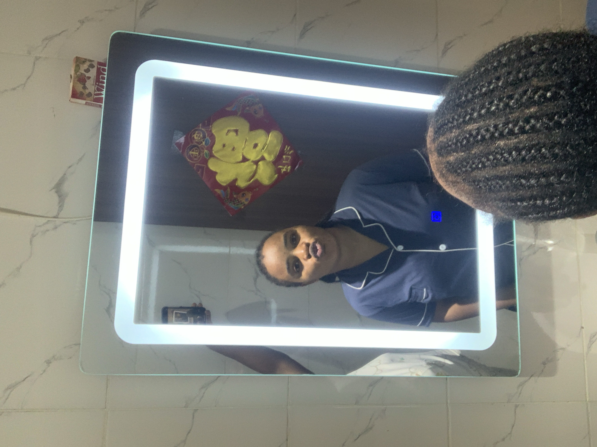 A woman with dark, braided hair is taking a selfie in a brightly lit, modern bathroom mirror. She is wearing blue pajamas with white trim and is making a puckered-mouth expression. In the reflection, a traditional Chinese New Year decoration with the character "Fu" (meaning luck or fortune) is visible on the wall, suggesting a festive atmosphere. Above the mirror, a small box labeled "Wind" with images of strawberries is placed on the tiled wall. The bathroom features marble-patterned tiles and a clean, contemporary aesthetic. The overall scene appears to be a personal moment captured in a private setting.