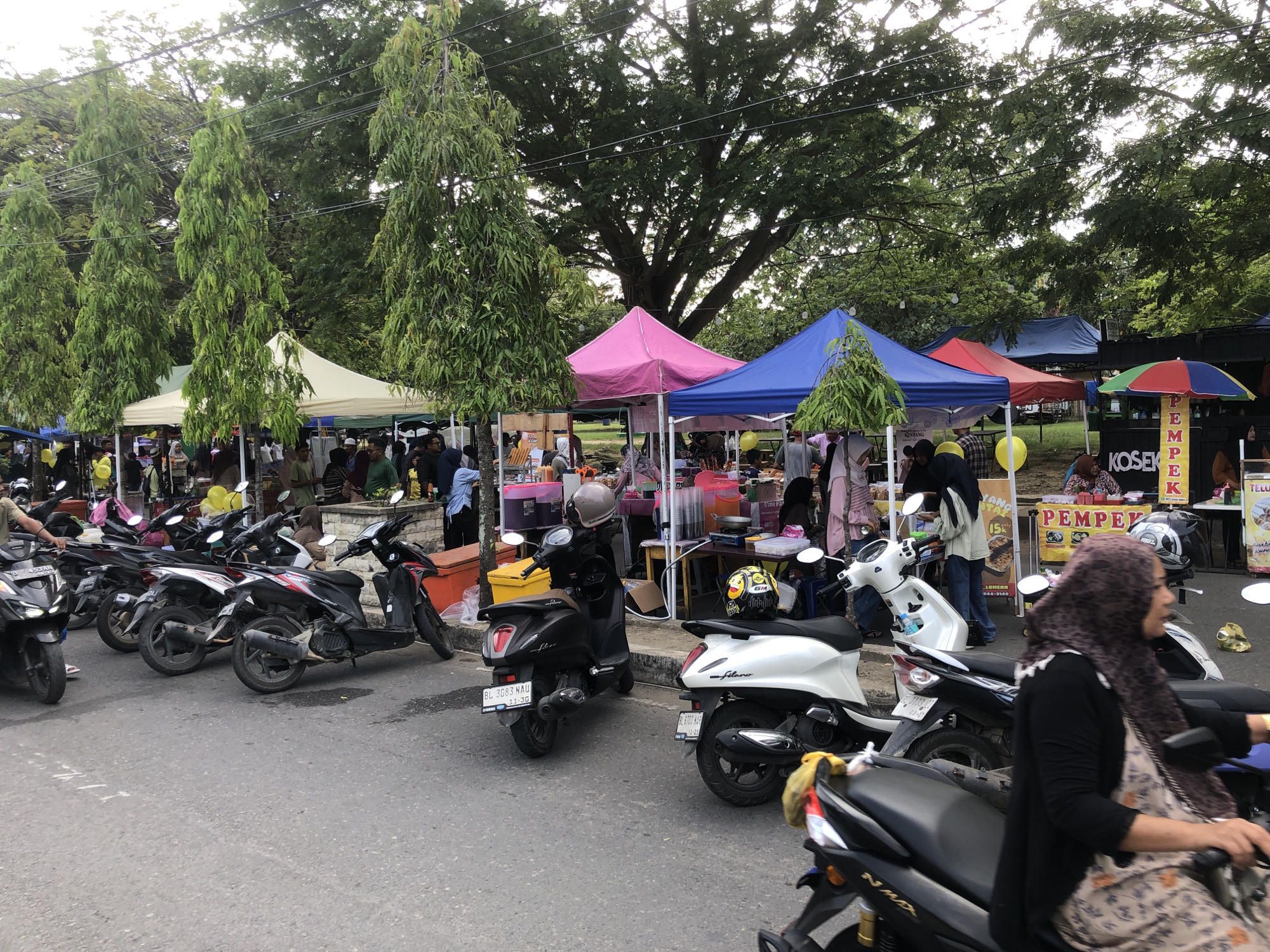 The image depicts a street scene in Lhokseumawe, Indonesia, lined with market stalls under colorful tents and a variety of parked motorbikes. The tents, in hues of cream, pink, blue, and red, suggest a bustling outdoor market. Vendors and customers are partially visible within and around the stalls, implying active commerce. A woman wearing a hijab is seated on a motorbike, possibly preparing to leave. Signage at a stall features the word "PEMPEK," indicating a local food offering, and "KOSEK," likely a vendor name. Banners advertise food offerings with "KENTANG" and "Umi Fa". Registration plates visible on some bikes suggest they are in active use, and trees in the background provide a natural backdrop. The street, partially filled with motorbikes, suggests a typical level of vehicular activity for a market area in Indonesia.