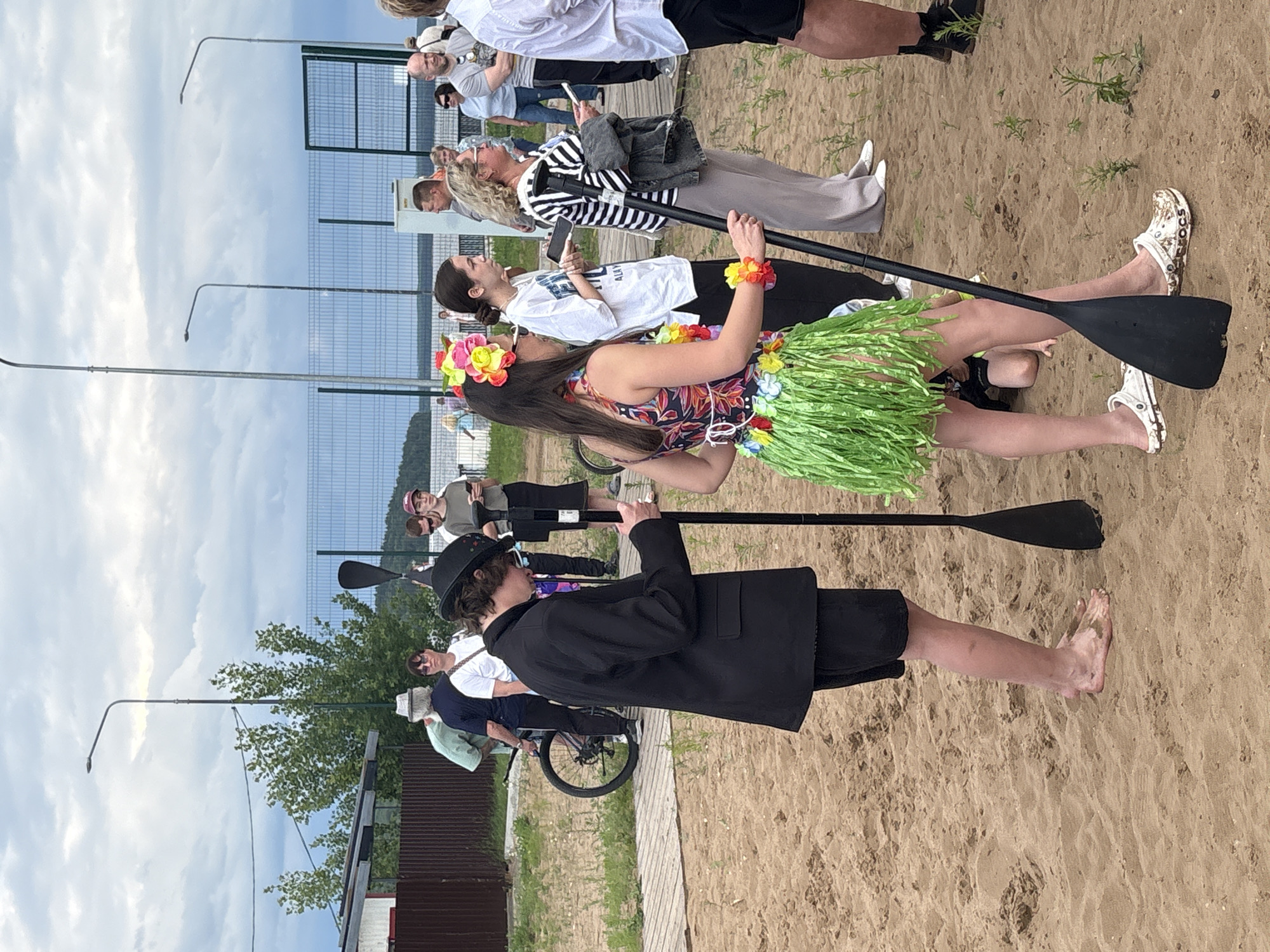 On a sandy embankment in the city of Chaikovsky, Russia, a group of people are gathered. In the foreground, a person dressed in a black suit and hat stands barefoot, holding a paddle. To their right, a person in a colorful Hawaiian-themed outfit, complete with a lei and grass skirt, also holds a paddle. Behind them, other individuals are visible, some holding phones and some appearing to be in conversation. The background shows a body of water, a fence, and streetlights, suggesting an outdoor event or gathering by the water. The environment appears to be a park or recreational area.