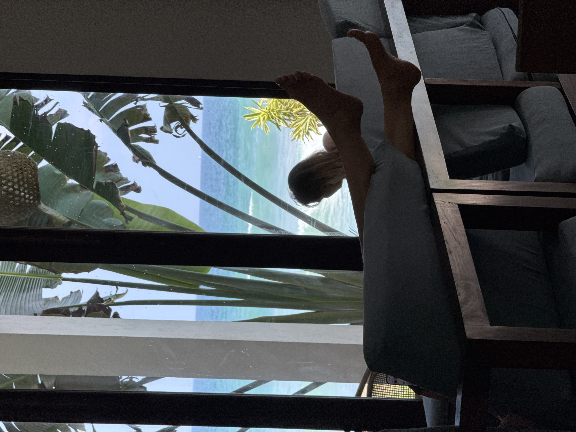 A person is lying on a sofa, legs raised towards a window. The view from the window shows a tropical beach with turquoise water, palm trees, and a sunny sky. The sofa is constructed with a dark wood frame and light gray cushions. The location is likely Goviyapana, Sri Lanka, based on the presence of tropical vegetation and beach scenery.