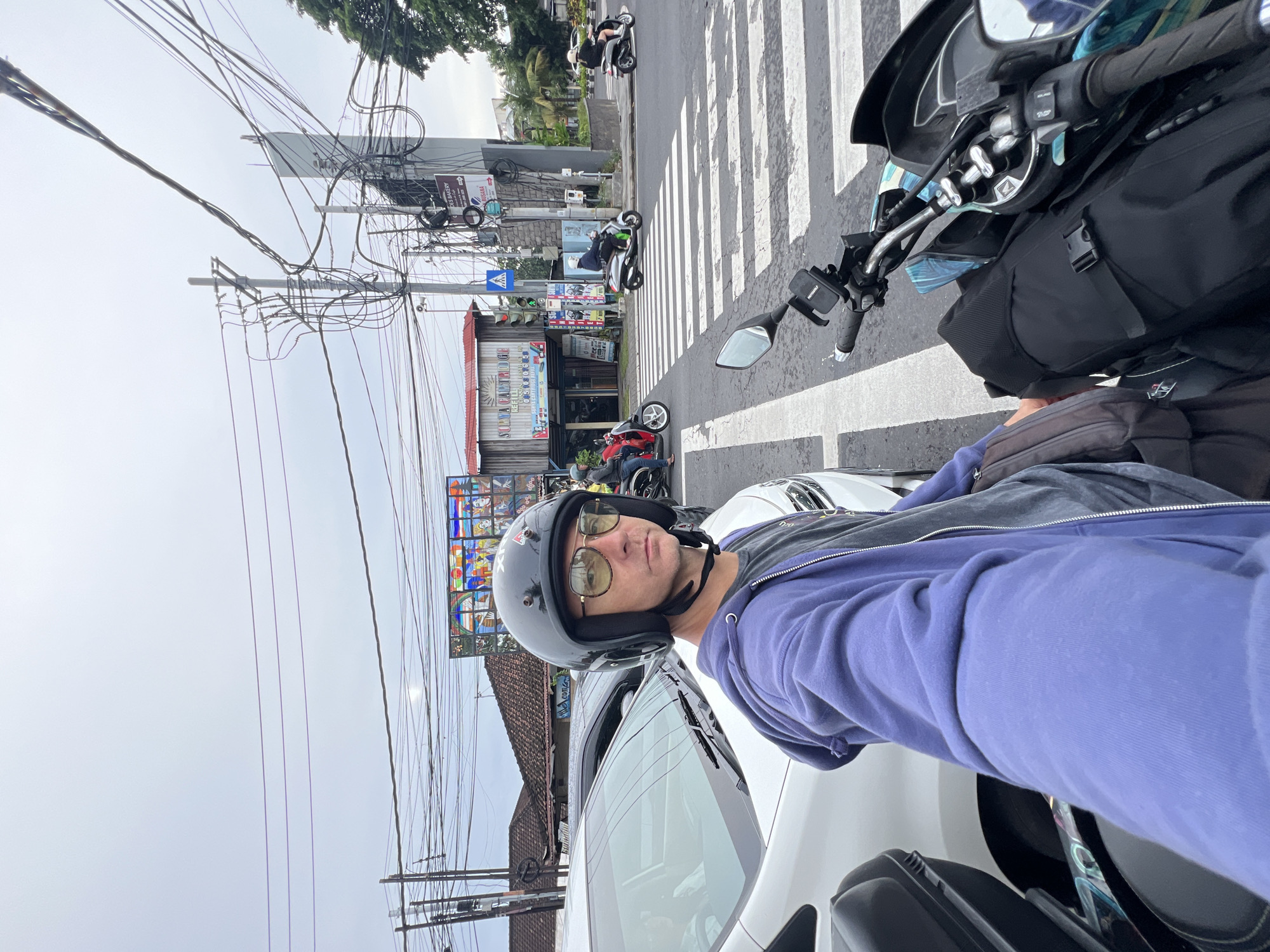 A man in a helmet and sunglasses is taking a selfie on a motorcycle in Denpasar, Indonesia. The background shows a street scene with utility poles, buildings, and other motorcycles. The foreground includes the man and the motorcycle he is on, which is stopped at a zebra crossing. Other vehicles and pedestrians can be seen in the distance. The location appears to be an urban area with commercial buildings and infrastructure elements typical of a city street. A white car is next to the motorcycle.