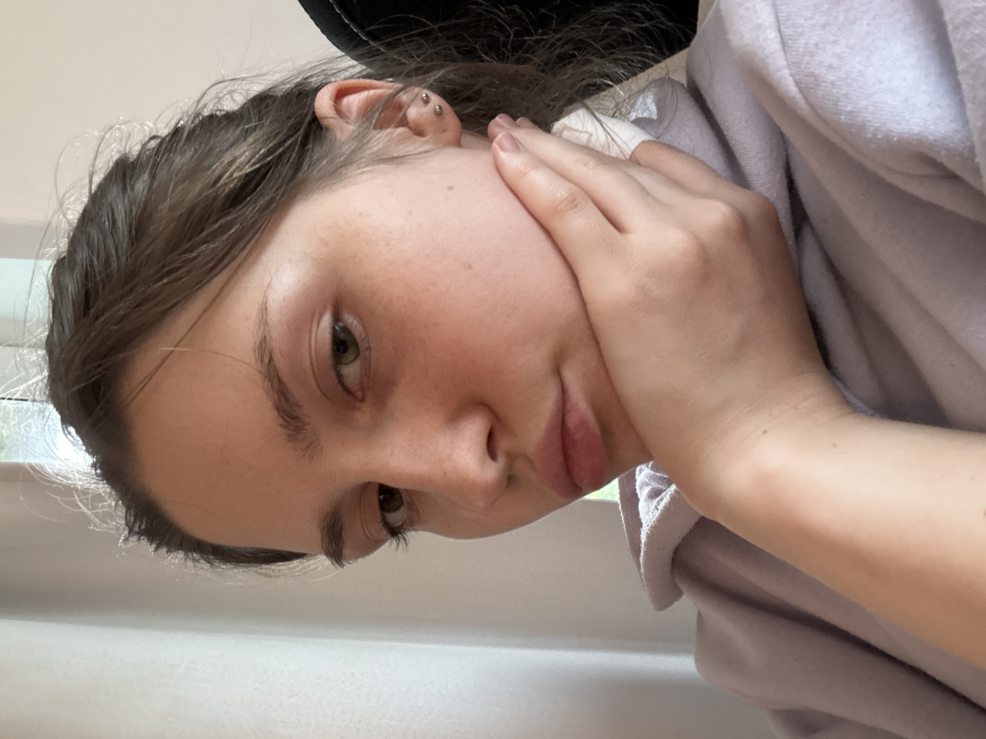 A close-up, angled portrait features a young person, likely female, with fair skin and light brown or hazel eyes, looking towards the viewer. Her head is tilted, and her left hand gently supports her left cheek. Her hair is dark brown and appears slightly textured, with lighter strands framing her face. She has subtle freckles visible on her cheek. Her left ear displays a double helix piercing with two metallic stud earrings. She is wearing a soft, light-colored garment, possibly a pale purple or off-white hoodie or sweater, visible around her neck and shoulder area on the right side of the frame. A dark object, possibly a hat or headphones, is partially visible above her head on the right. The background is softly blurred, showing bright, light-colored surfaces that suggest an indoor setting with natural light illuminating her from the left. Her expression is calm and somewhat pensive, with her lips slightly parted in a subtle pout. There is no visible text in the image. While the location context is provided as Saint Petersburg, Russia, no specific visual cues within the image itself confirm this.