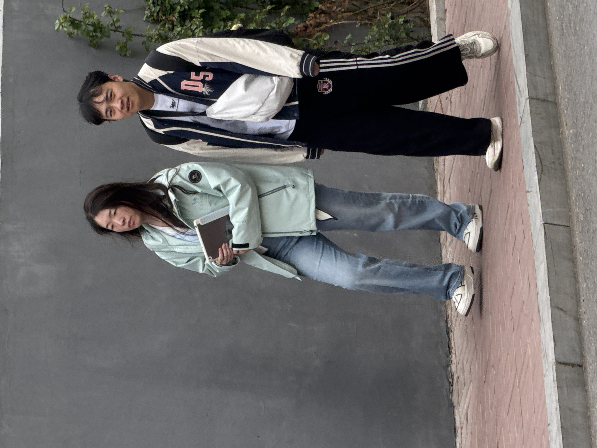 Two people are standing on a brick walkway next to a gray wall, which is located in Zhongtun, China. A person wearing a black and white jacket with the number "05" on it, paired with black Adidas track pants and white sneakers is to the left. The second person, on the right, is wearing a mint green jacket, blue jeans, and white sneakers. They are holding a tablet. Behind the first person is some greenery.