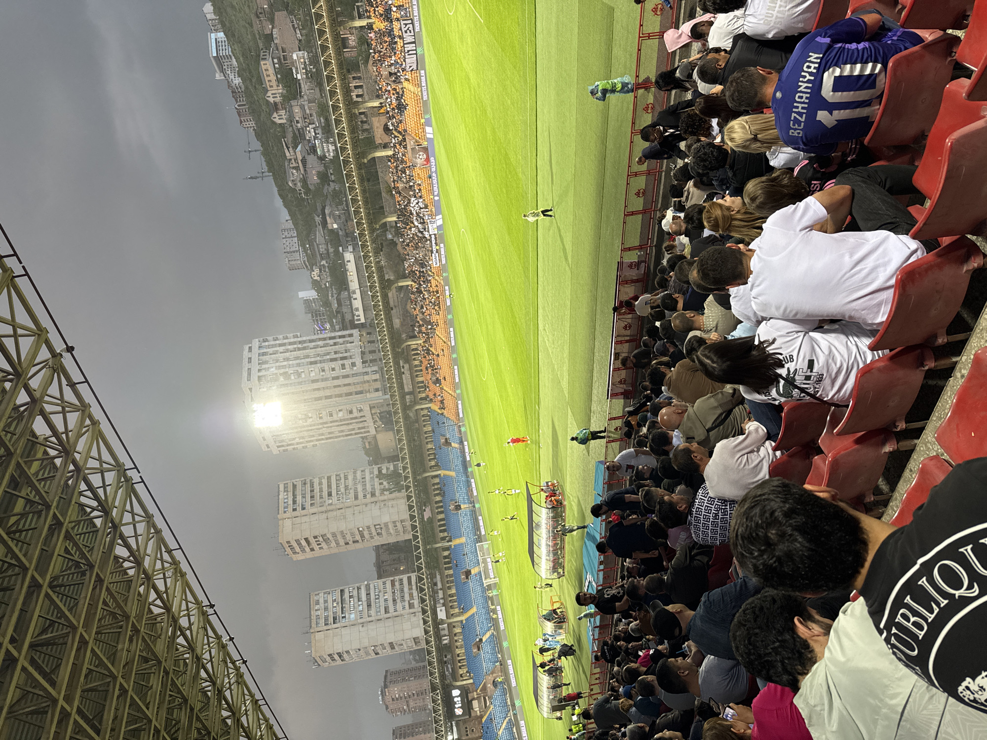 Here's a detailed description of the image:
**Scene:** A wide-angle shot taken from a stadium during what appears to be a soccer (football) match.
**Location:** The stadium is located in Yerevan, Armenia. The OCR "ՀԱՅԱՍՏԱՆԻ ԳԱՎԱԹ," which translates to "Armenian Cup" is visible.
**People:** The stadium is densely populated with spectators. Many people are seated in the red stadium chairs, closely packed together. The crowd is diverse in age and attire. Some are wearing jerseys, casual shirts, and jackets.
**Subjects and Objects:**
* Soccer field: A brightly illuminated green field is central to the image.
* Players: Several players are visible on the field, indicating active gameplay.
* Cityscape: A dense urban environment is visible beyond the stadium, with high-rise buildings and residential areas.
* Stadium Structure: The stadium's steel framework is visible on the left side of the image.
* Banners: Banners with text related to the game are visible along one side of the field.
**Actions and Interactions:** The primary action is the ongoing soccer match. The spectators are watching the game, presumably reacting to the events unfolding on the field.
**Environment:** The sky is overcast, suggesting either dusk or a cloudy day. The stadium lights are on, indicating that the game is either being played during the evening or under low-light conditions.