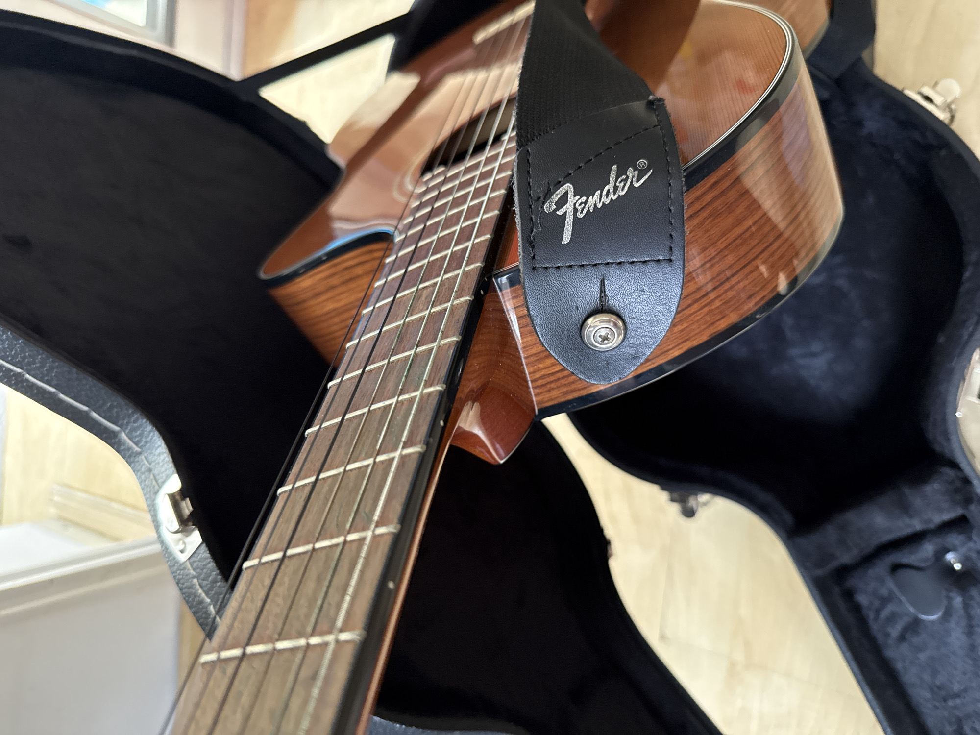 This close-up, angled photograph captures an acoustic guitar partially resting within its open, black hardshell case. The guitar features a glossy, light-brown wooden body with a visible grain, and a wide fretboard equipped with frets and strings, suggesting it is a classical or nylon-string model.
A prominent black leather guitar strap is attached to the guitar's body. The strap is clearly branded with the "Fender" logo in silver or white script, secured by a visible metallic snap button. The guitar's shiny surface reflects ambient light, highlighting the wood's texture and the instrument's contours.
The interior of the black guitar case is lined with a soft, plush material, creating a dark, protective cradle for the instrument. A small accessory compartment or support within the case is visible towards the lower right. In the background, bright light streams from what appears to be a window, indicating an indoor, daytime setting. A light-colored surface, possibly a floor or wall, is also seen. The scene conveys a sense of the guitar being stored, prepared for playing, or recently put away. The location context for this image is Blagoevgrad, Bulgaria.